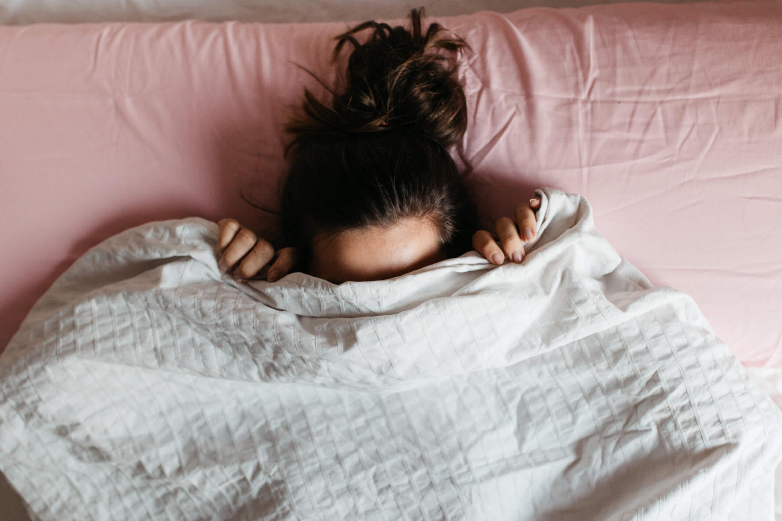 Verspielte junge Frau versteckt ihr Gesicht unter der Decke, während sie in einem gemütlichen Bett auf einem weißen