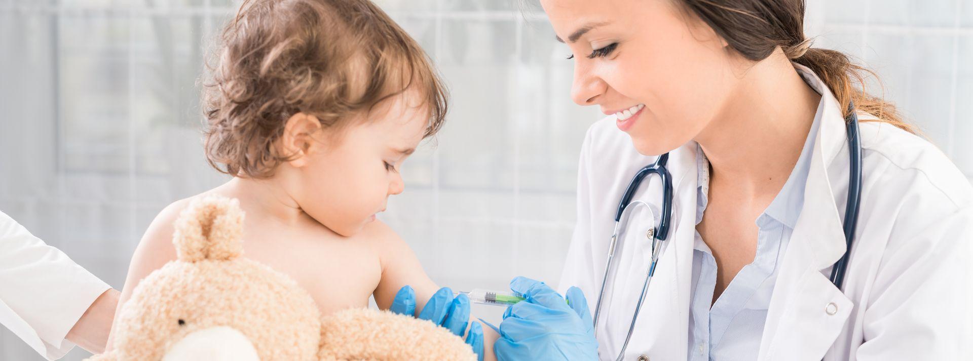 Kinderarzt der jungen Frau führt eine Impfung eines kleinen Mädchens durch. Das Mädchen hält ein Maskottchen.