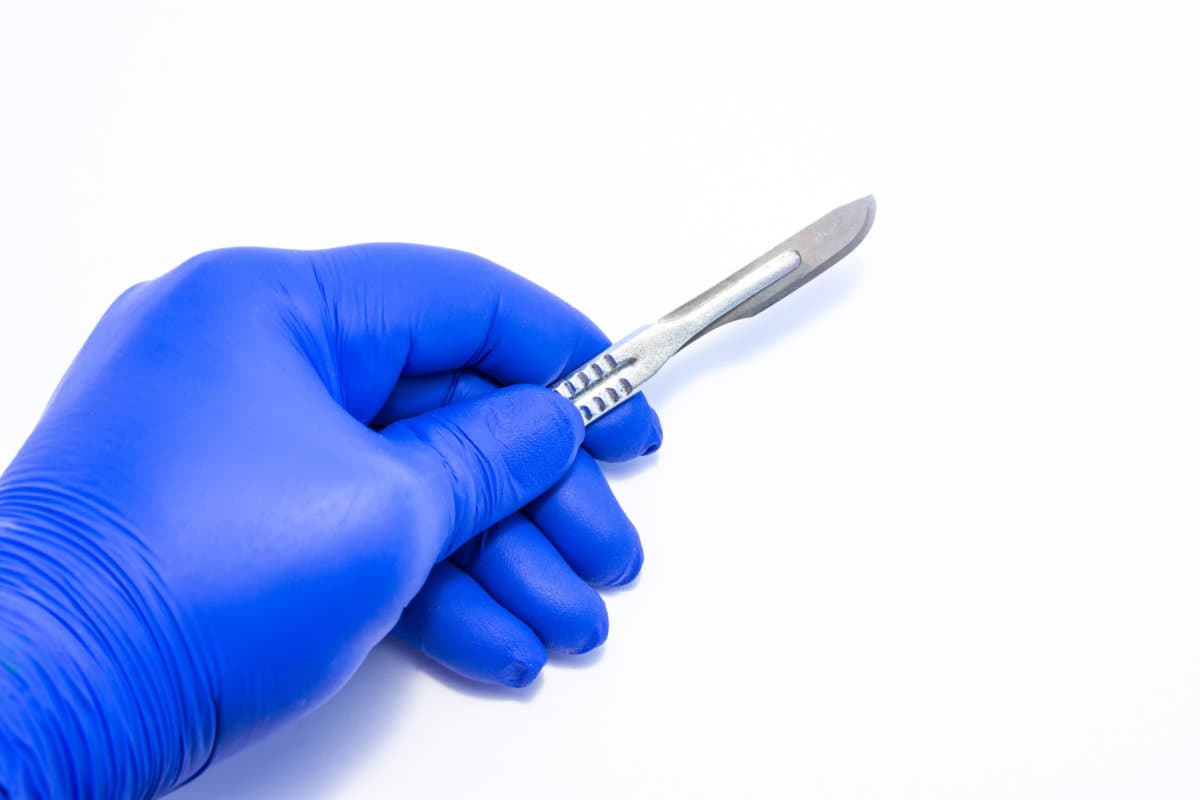 Concept photo surgery and surgical treatment, operations, procedures. Doctor holds in his hand, dressed in blue medical glove, metal scalpel with removable blade on white homogeneous background