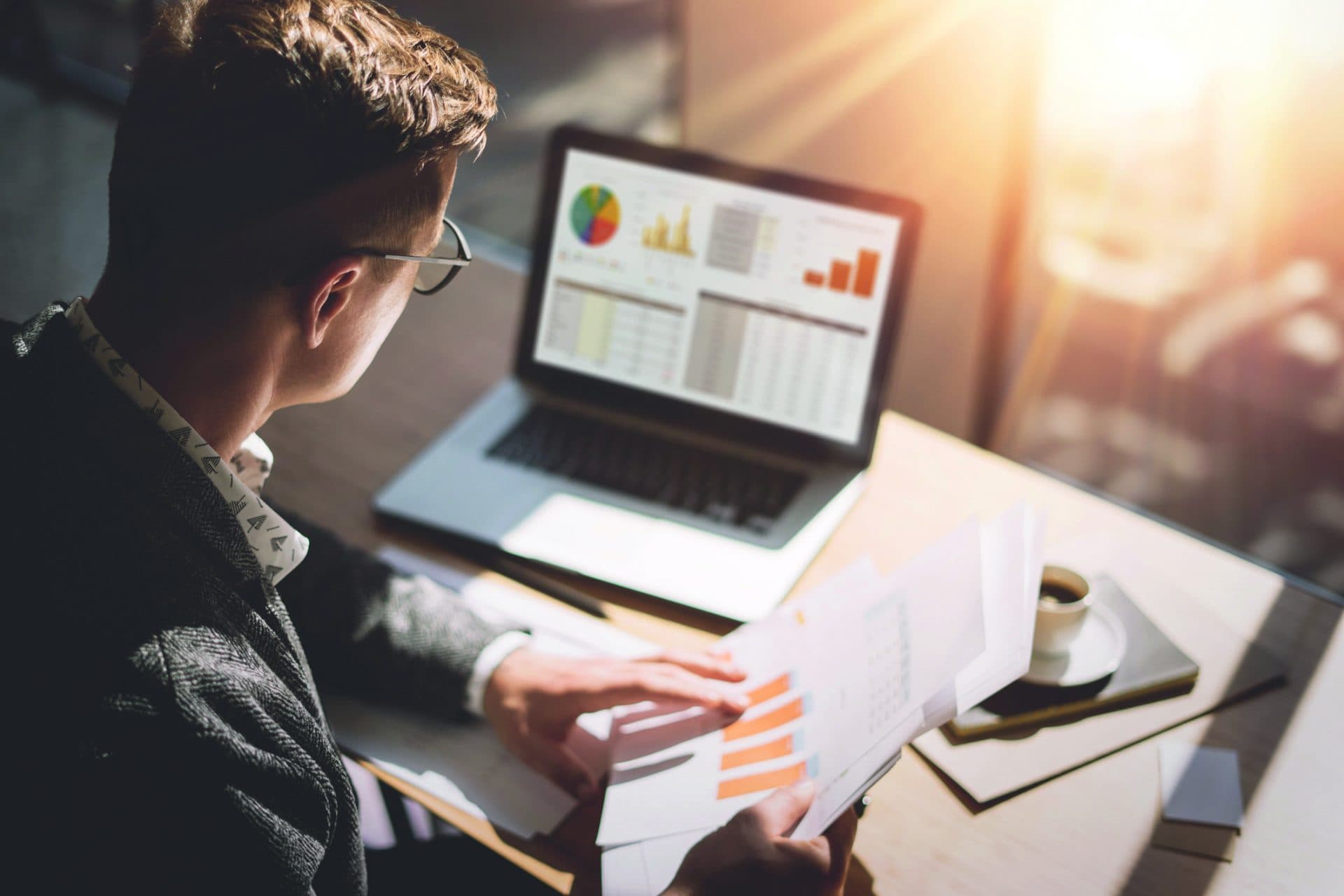 Junger Finanzmarktanalyst in Brillen, die am sonnigen Büro auf Laptop arbeiten, während sie am Holztisch sitzen. Geschäftsmann analysieren Dokument in seinen Händen. Graphen und Diagramm auf Notizbuchbildschirm. Unscharf.