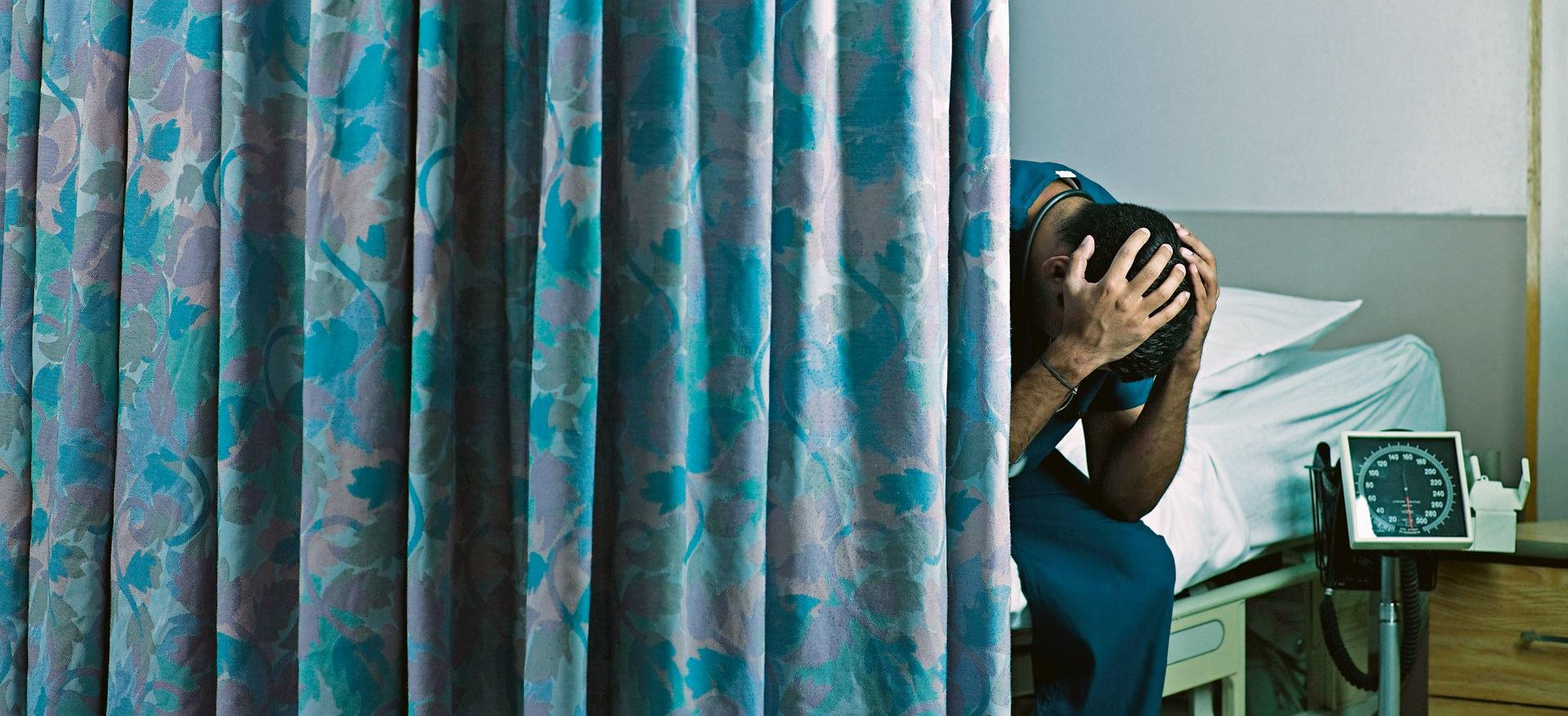 Foto eines halb von einem Vorhang verdeckten Mannes, der, auf einem Bett sitzend, sein Gesicht in den Händen vergräbt.