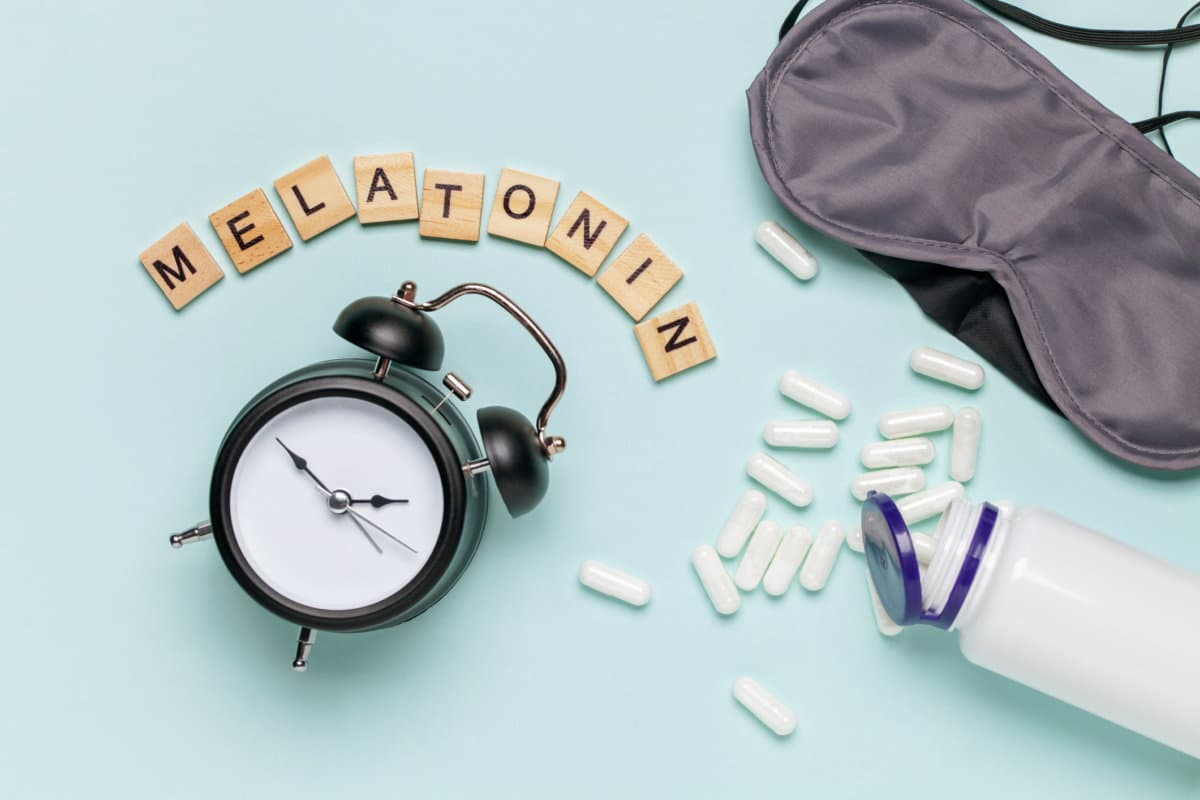 Weiße Flasche und Pillen, Buchstaben Melatonin, Wecker und Schlafmaske auf hellgrauem Hintergrund.