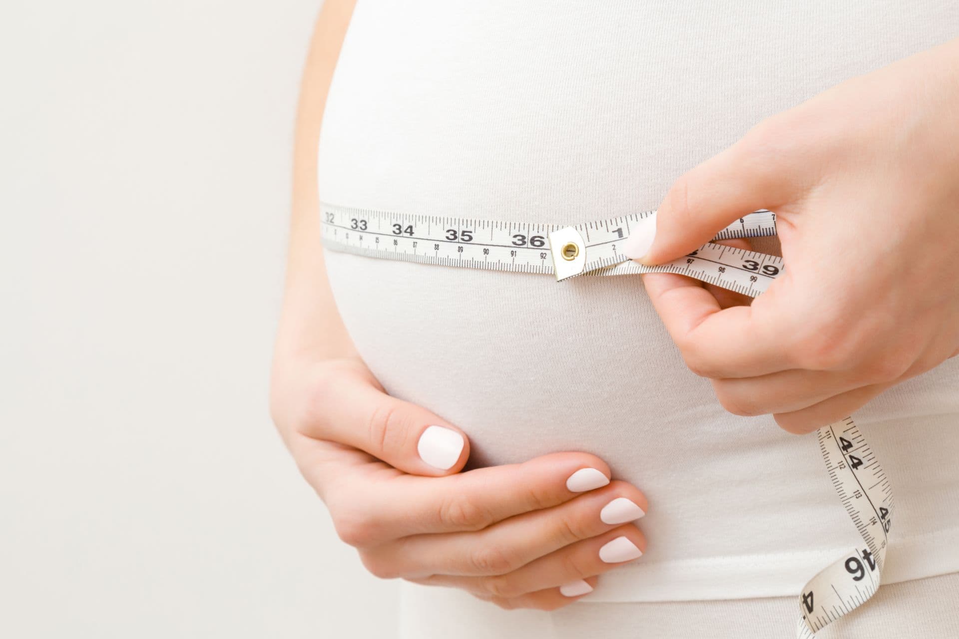 Junge Frau Hand, die dicken Bauch auf grauem Hintergrund misst. Schwangerschaftszeit - 20 Wochen. Baby Erwartung. Nahansicht. Seitenansicht.