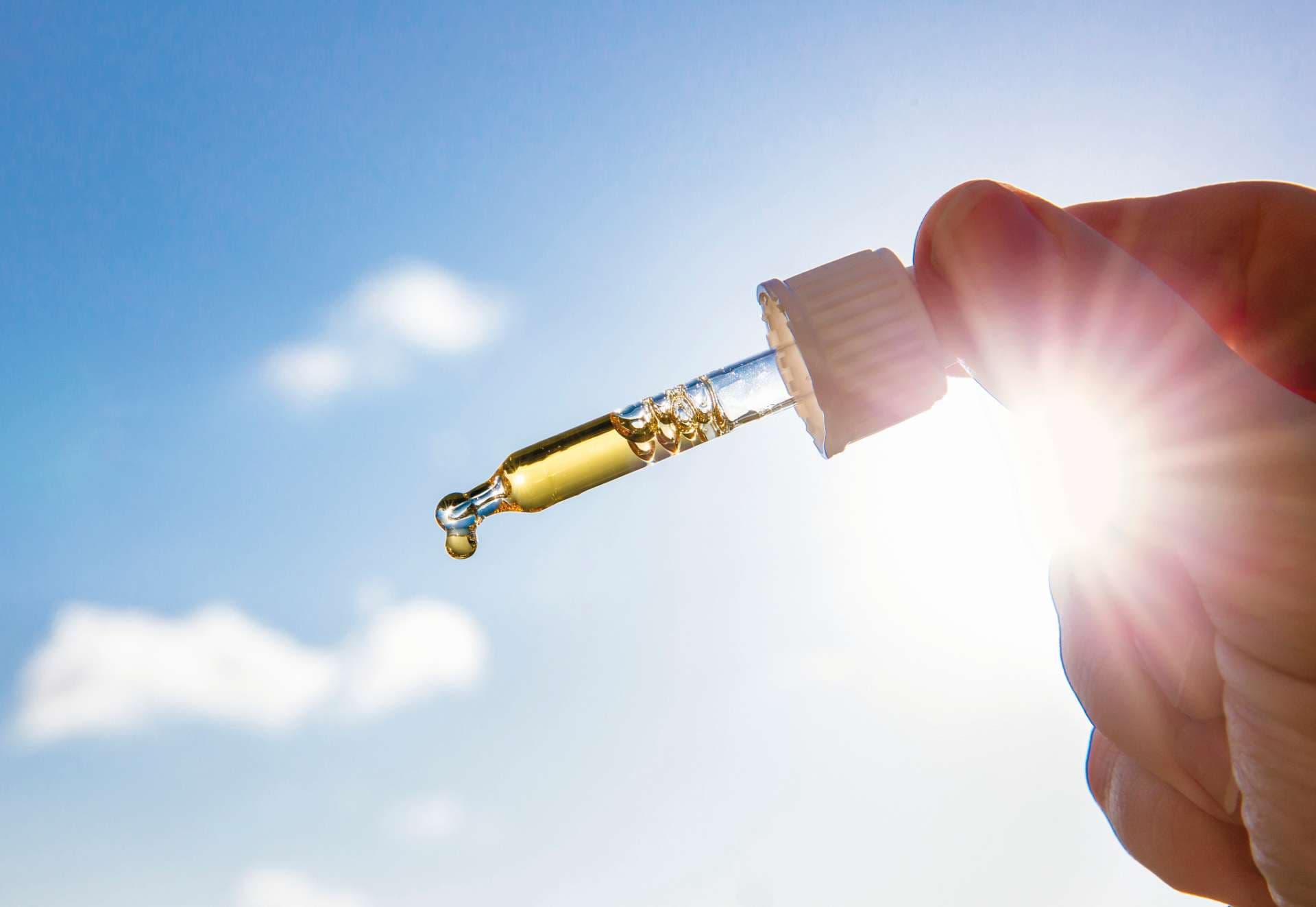 Hand, die Pipette mit schönem goldenem flüssigem D-Vitamin gegen Sonne und blauen Himmel an einem sonnigen Tag hält. Vitamin D hält Sie bei Sonnenmangel im Winter gesund, Kurkonzept.