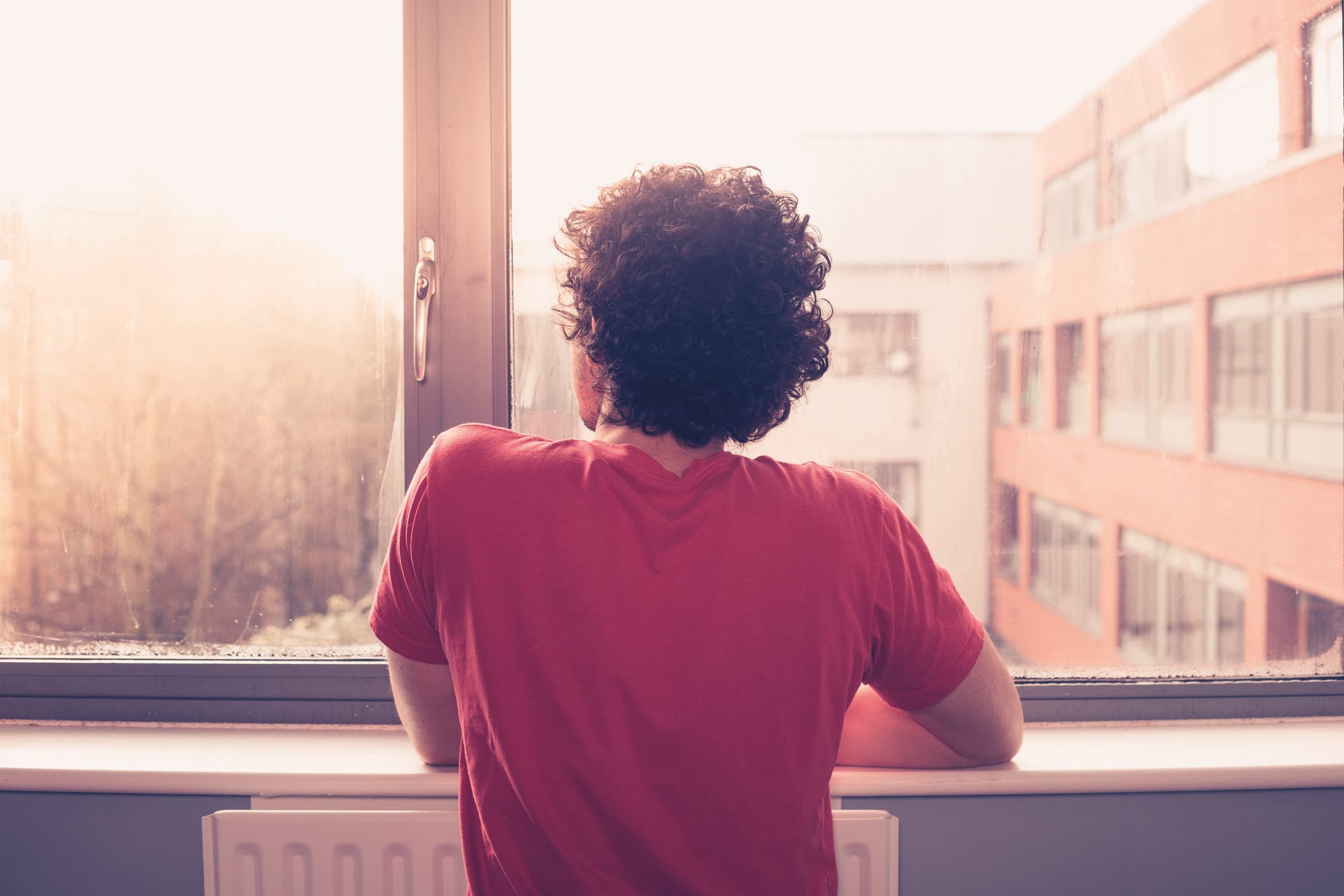 Junger Mann steht am Fensterbrett und schaut auf den Sonnenuntergang