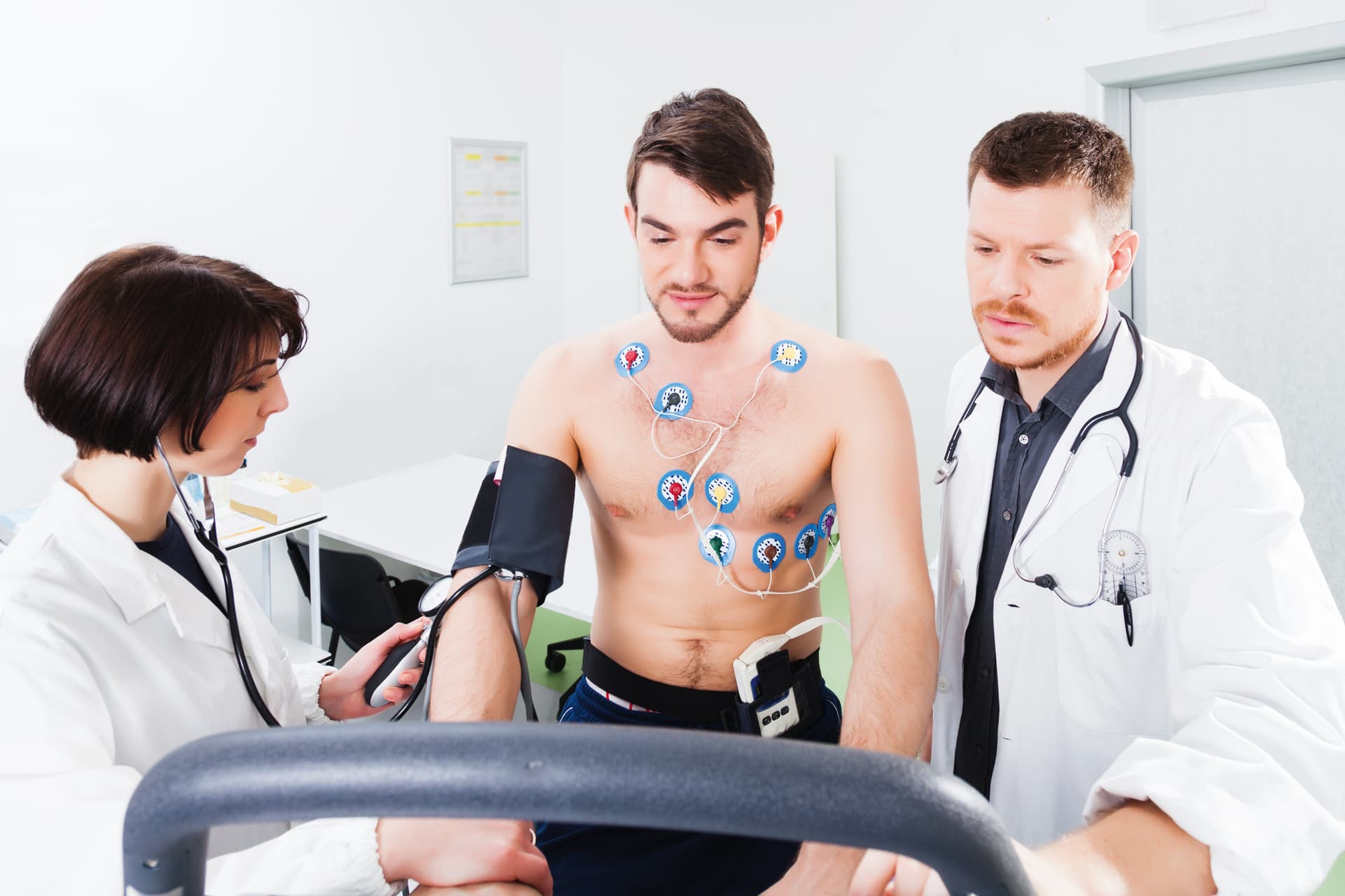 Ein Mann steht auf einem Laufband und jeweils ein Arzt steht links und rechts daneben. Der Mann ist oberkörperfrei und an Elektroden gesteckt.