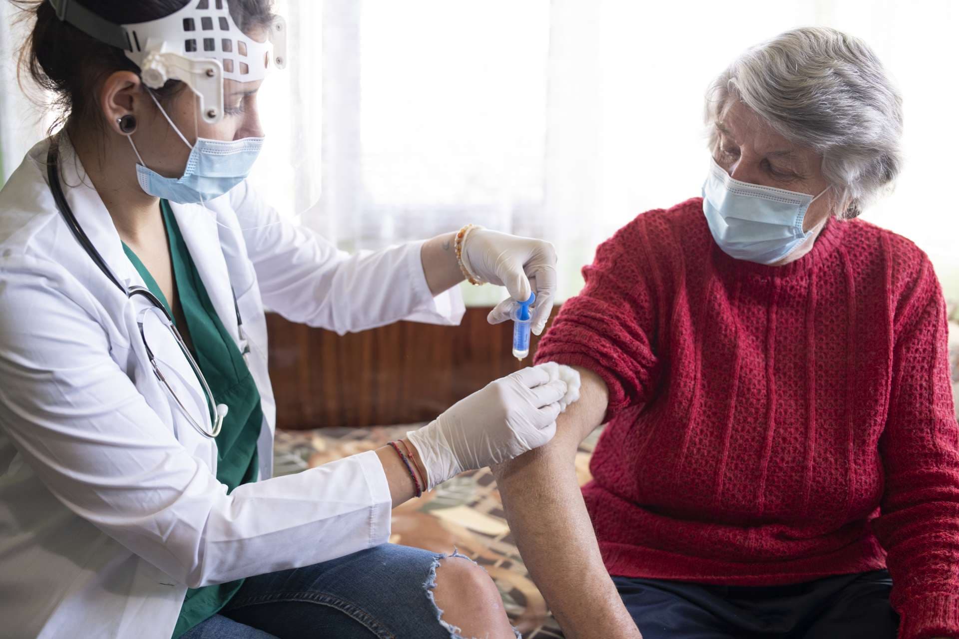 Weibliche Gesundheitsperson, die der älteren Frau in ihrem Haus Impfstoff injiziert.