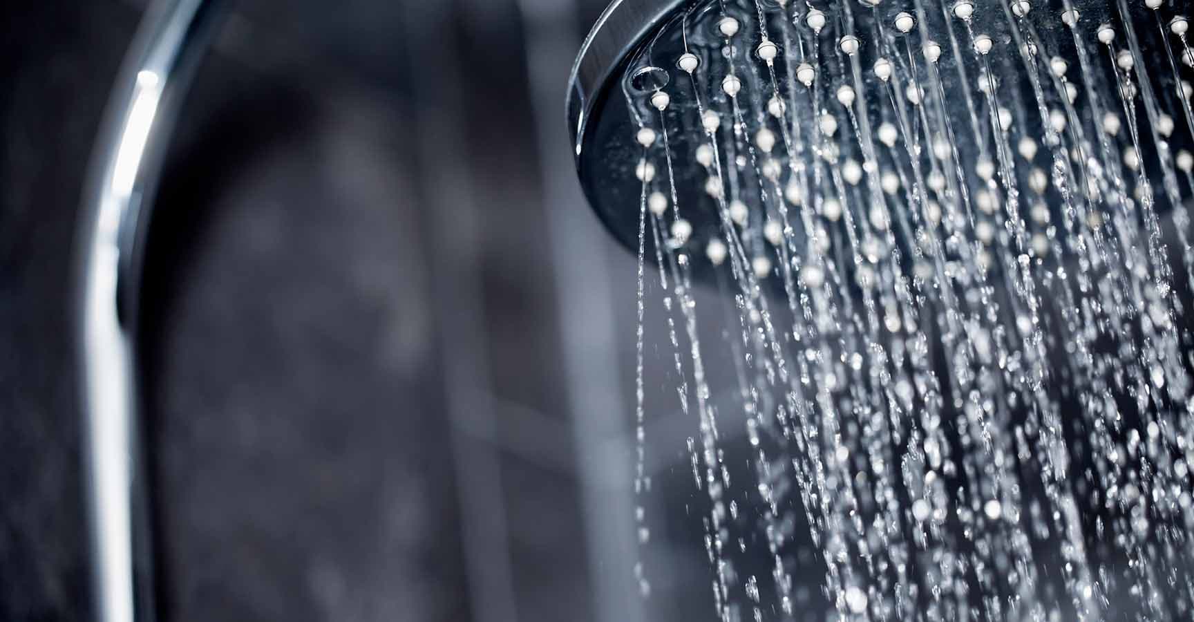 Close up view of large shower head sprinkling water with black tiles in the background.