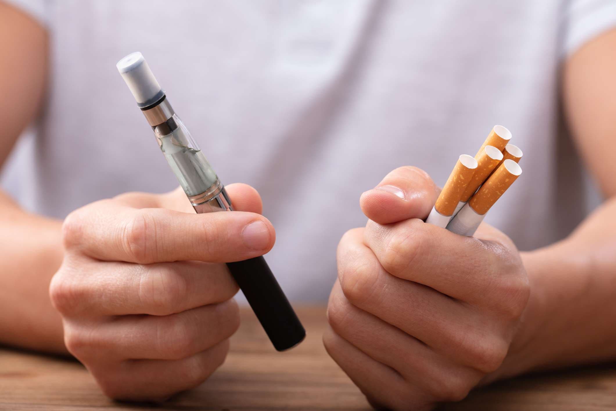 Man Holding Vape And Tobacco Cigarette Over Desk