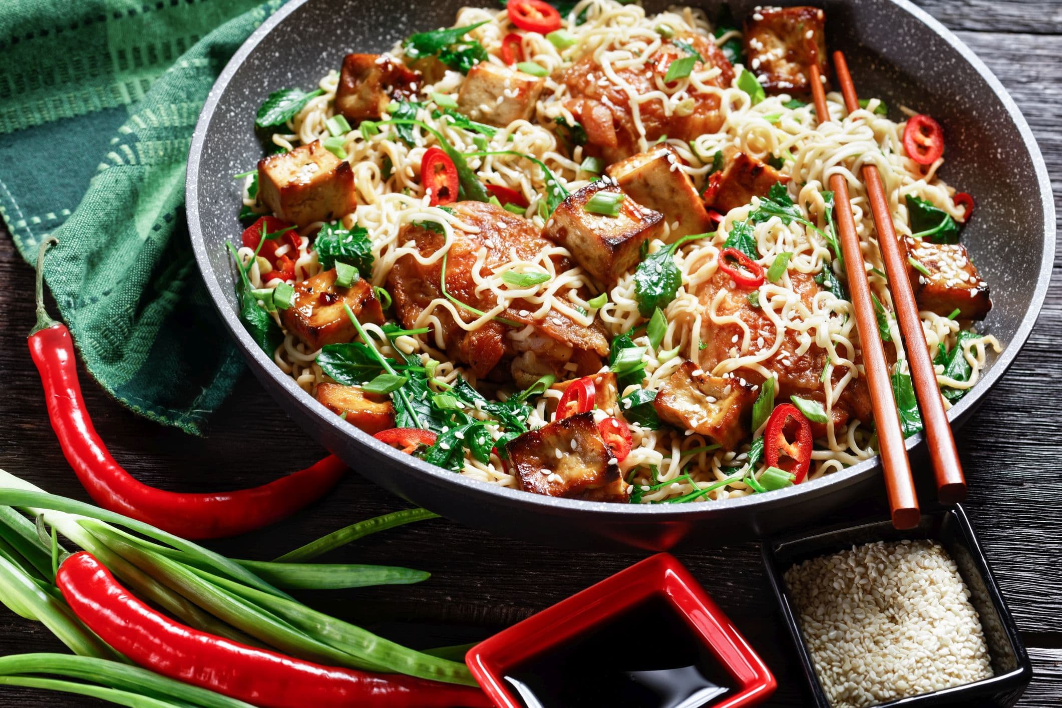 Süß-saurer Tofu, Hähnchenschenkel, chinesische Woknudeln, verwelkter Grünkohl, Sesam, roter Chili, Frühlingszwiebeln, serviert auf einer Pfanne mit Stäbchen auf dunklem Holzhintergrund, Draufsicht, Nahaufnahme