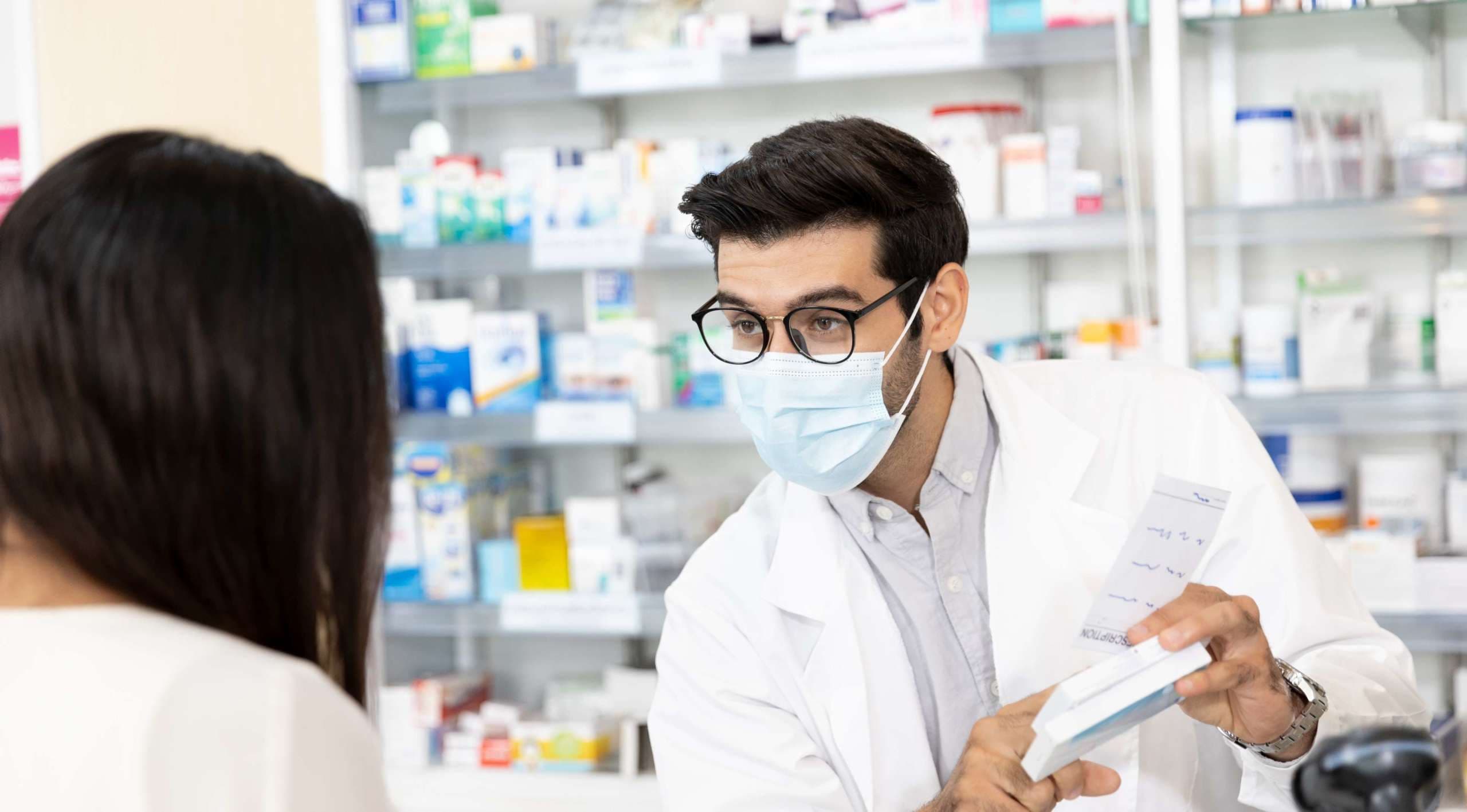 Apotheker, der eine hygienische Schutzmaske trägt und in der modernen Apotheke Medikamentenempfehlungen gibt