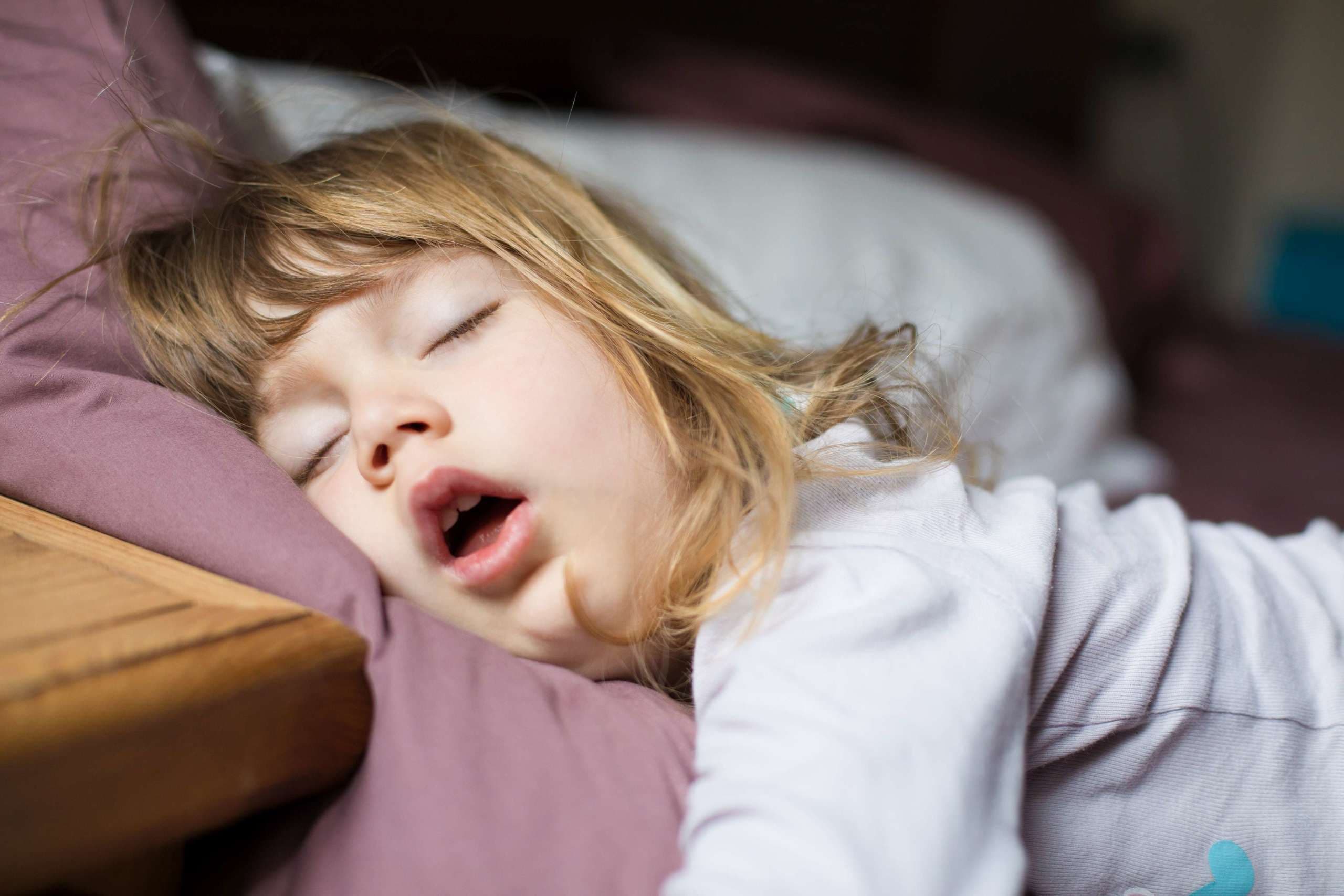 lustiger Gesichtsausdruck mit offenem Mund eines blonden, kaukasischen dreijährigen Kindes, das auf einem Kingsize-Bett schläft
