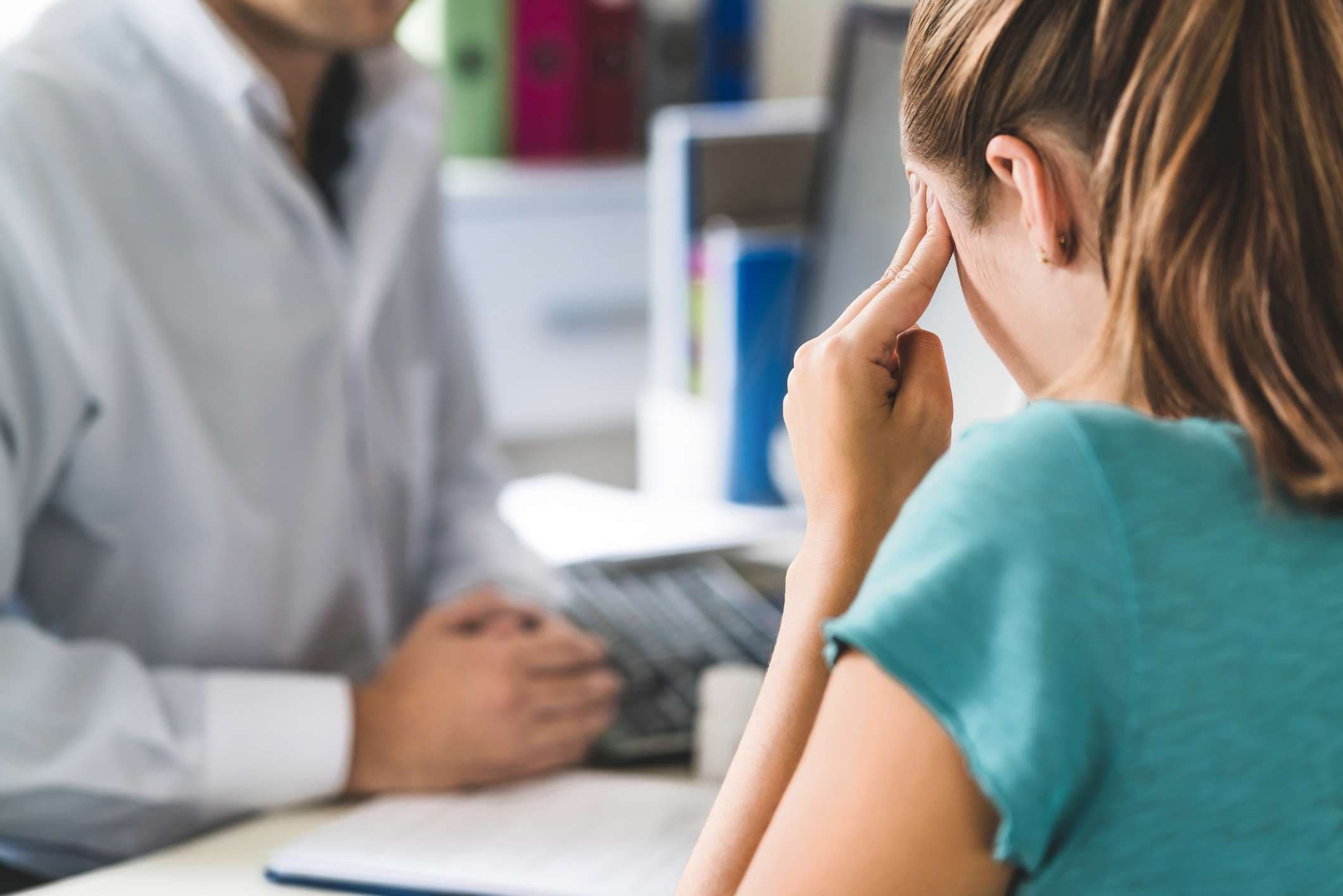 Frau, die unter starken Kopfschmerzen oder Migräne leidet. Termin mit Arzt im Büroraum. Kranke und unwohle Dame mit Stress, Trauma oder Burnout. Junge Patientin mit Schmerzen, die den Kopf mit den Händen hält.