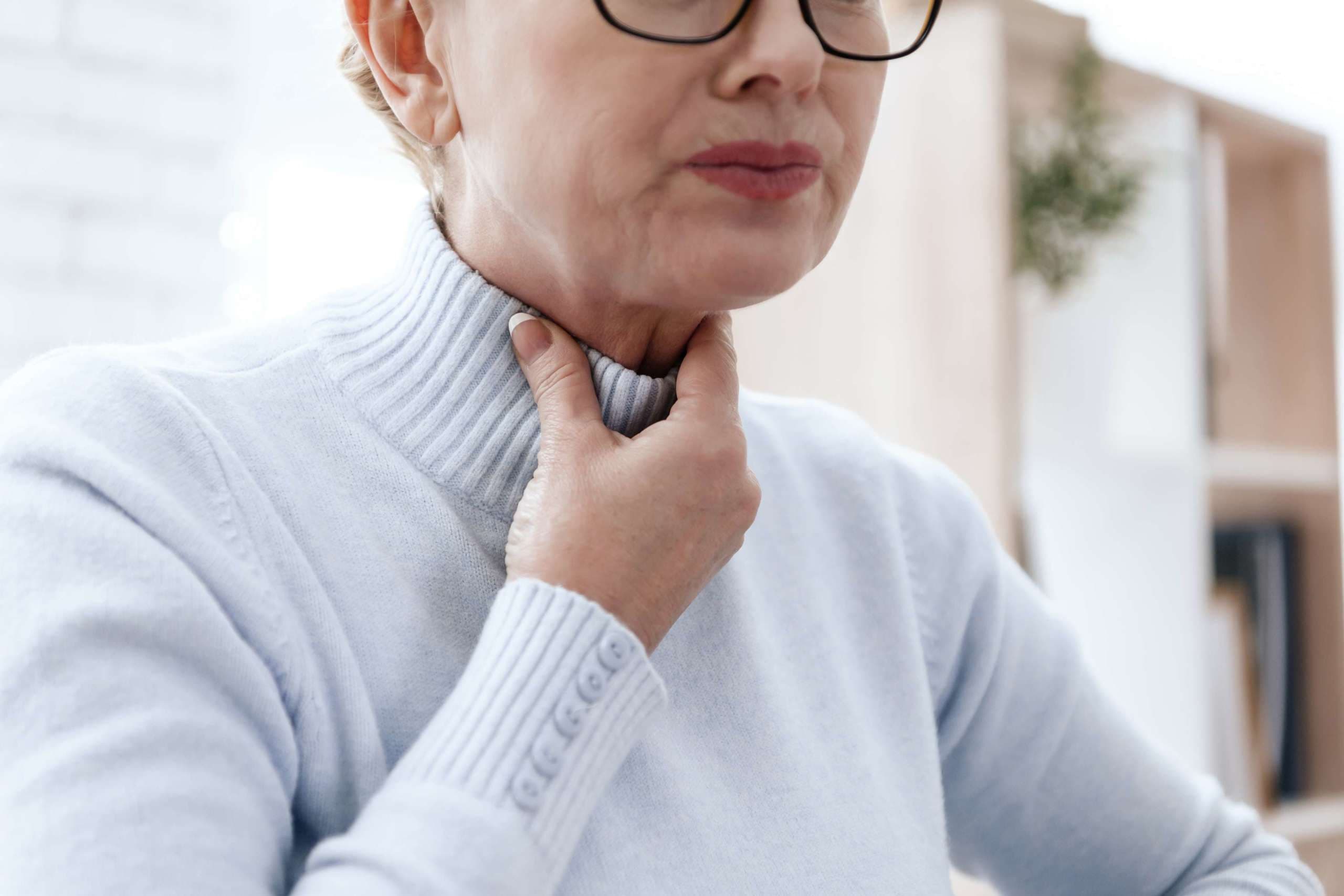 Eine Frau hat Halsschmerzen. Sie hat keine Stimme. Eine Grimasse auf seinem Gesicht. Sie hält ihre Hände an ihren Hals.