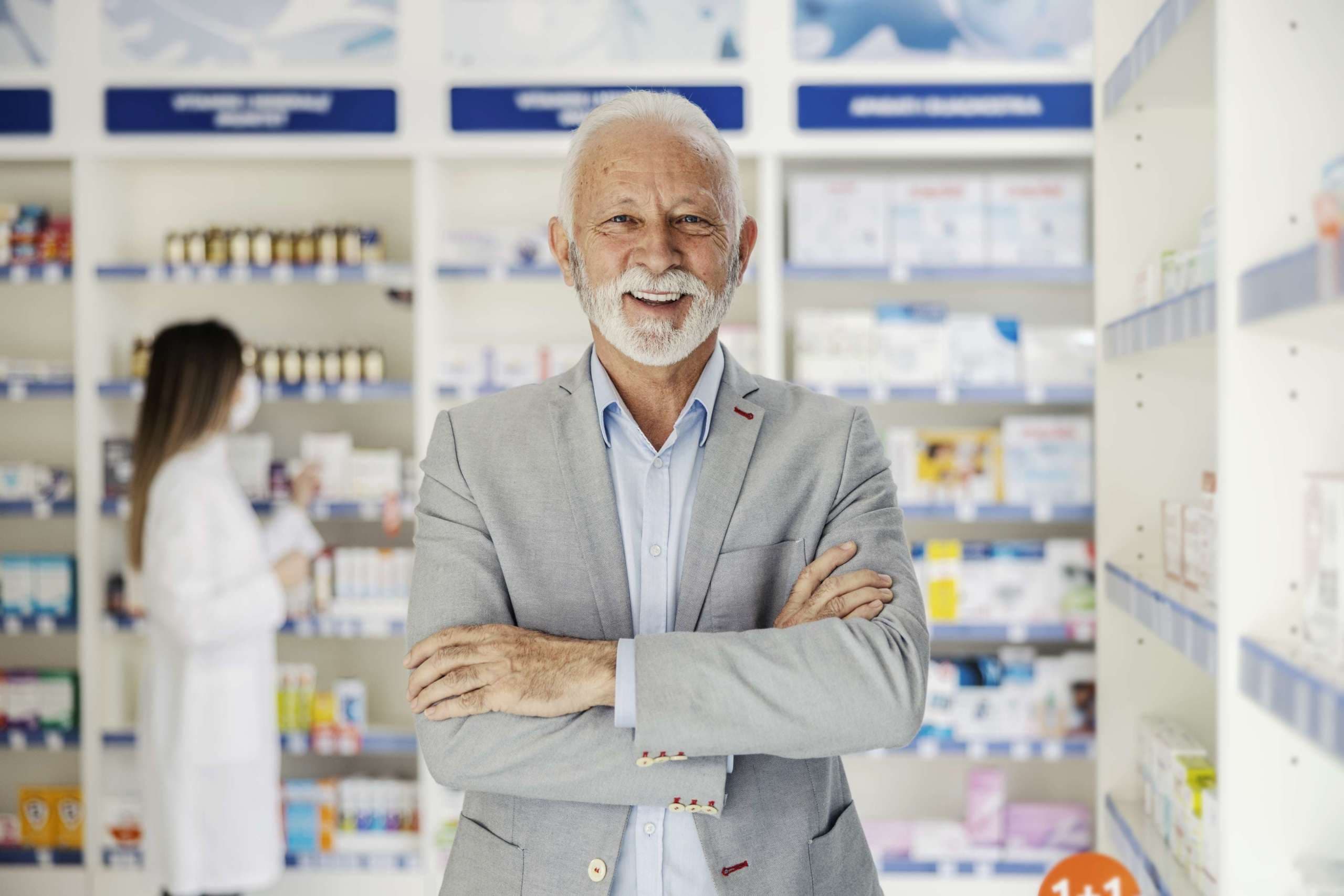 Reifer Mann mit verschränkten Armen steht in einer Apotheke. Porträt eines Mannes, der in einem eleganten Anzug in einer Apotheke arbeitet, nimmt eine positive Einstellung gegenüber Patienten und Kunden ein. Frau im Hintergrund verschwommen