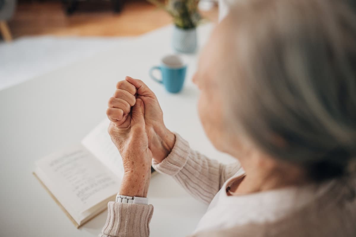 Eine Frau, eine alte Frau, hält ihre arthritischen Hände, während sie zu Hause sitzt und ein Buch liest.