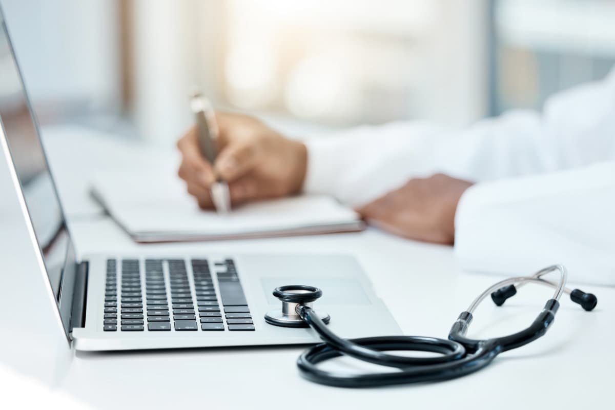 Laptop, stethoscope and doctor writing in notebook for research planning or medical tech innovation in hospital office. Healthcare medic worker, research strategy book notes and online communication