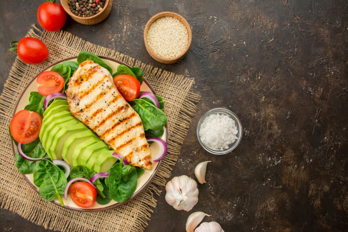 Gegrillte Hühnerbrust und frischer Gemüsesalat mit Spinatblättern, Avocado und Tomaten auf dunklem Hintergrund.