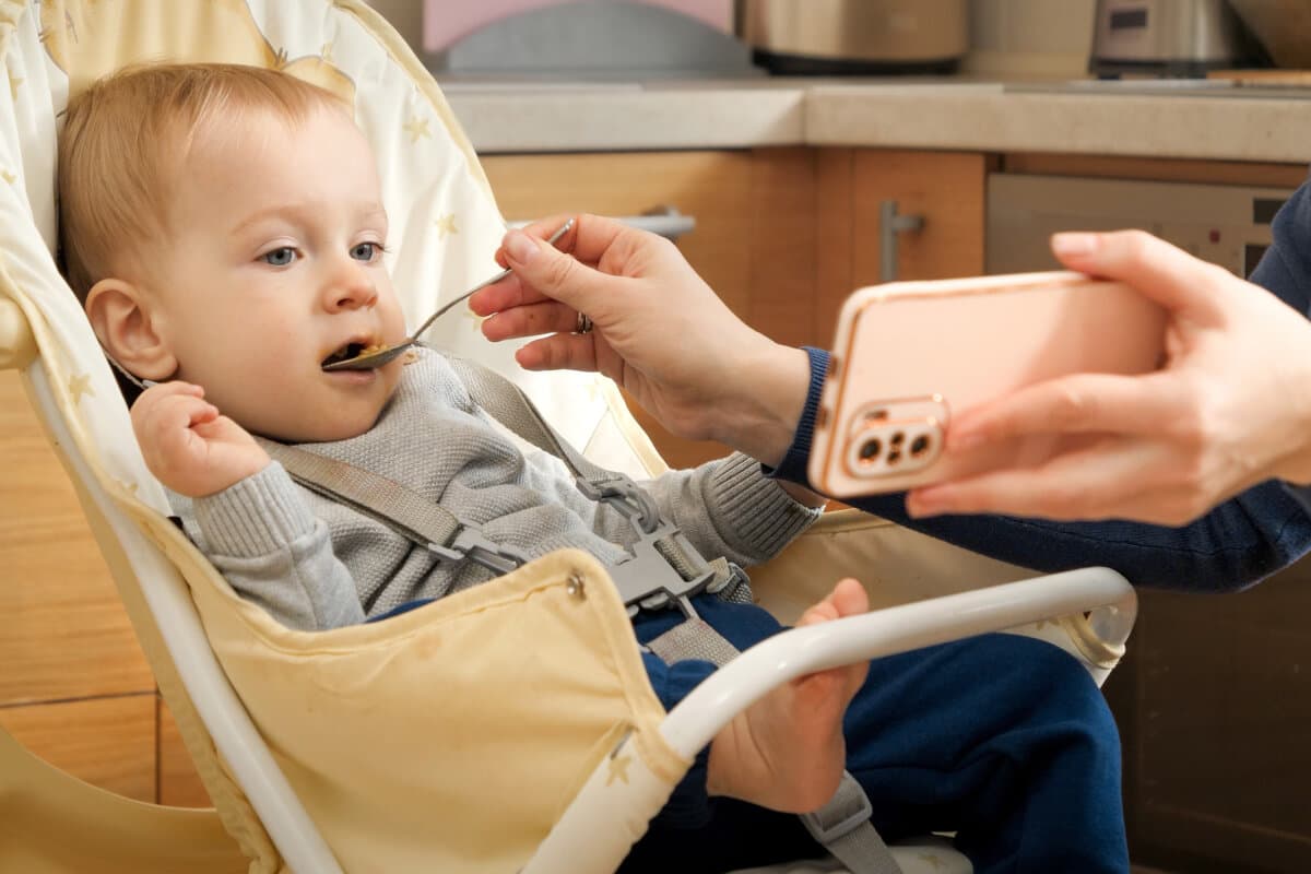 Mutter zeigt Video auf Smartphone, während sie ihr Baby im Hochstuhl füttert.