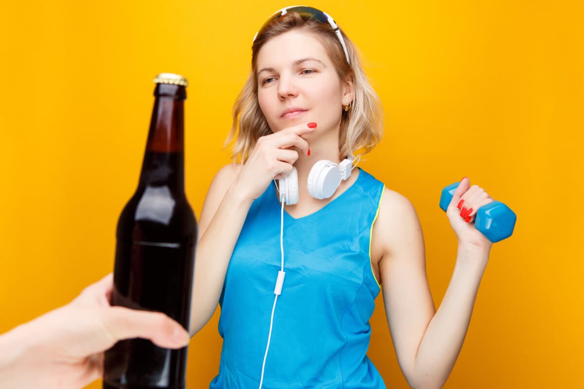 Sportliches blondes Mädchen mit Kopfhörern und Hantel in der Hand, vor der Hand eine Flasche Bier.