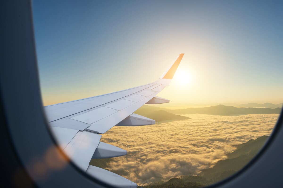 Flugzeug fliegt tief über nebligen Bergen, Blick aus dem Flugzeugfenster auf die Tragfläche und den Sonnenaufgang am Himmel