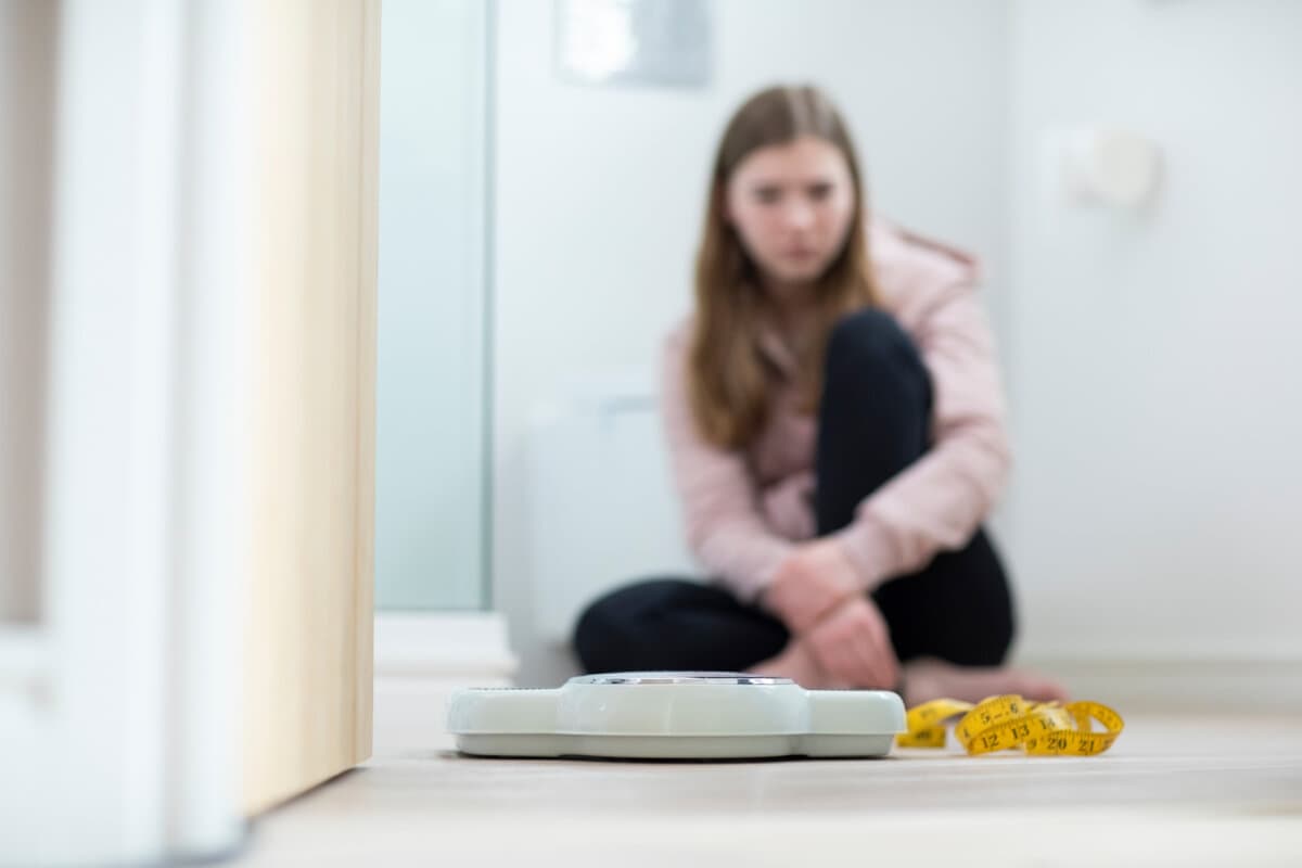 Unglückliches Teenager-Mädchen sitzt im Badezimmer und schaut auf Waage und Maßband