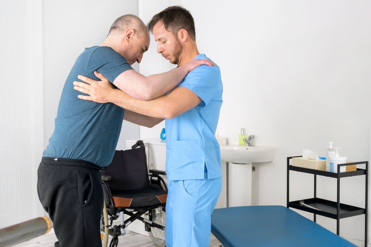 Männlicher Physiotherapeut hilft einem behinderten Patienten im Rollstuhl beim Aufstehen im Rehabilitationskrankenhaus.