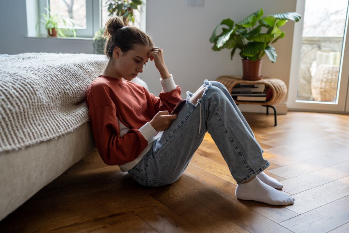Upset teen girl sitting on floor near bed using smartphone at home, teenagers and gadget addiction