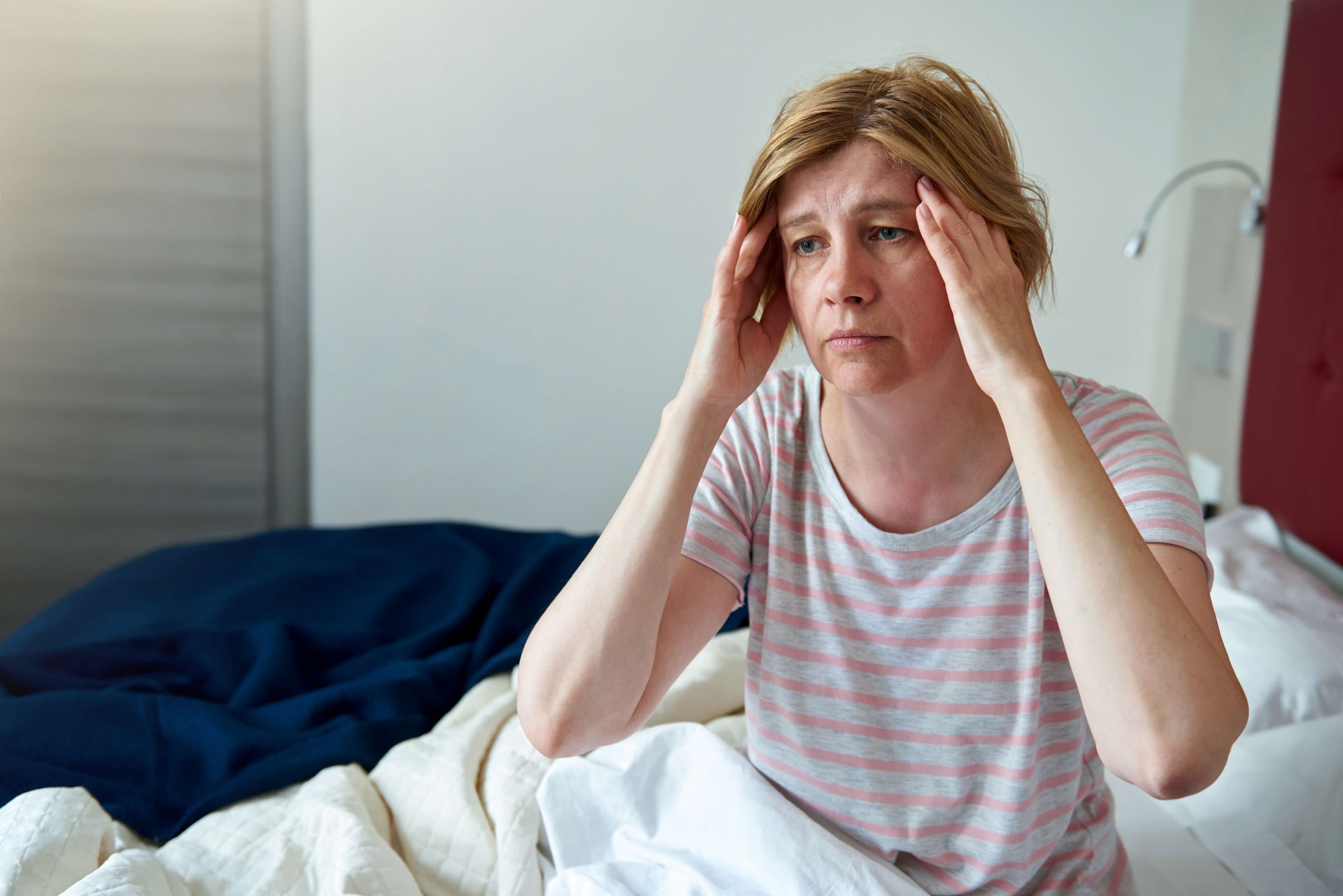 A woman, 50 years old, is sitting on the bed, holding her head. Health problems, headache, menopause, insomnia.
