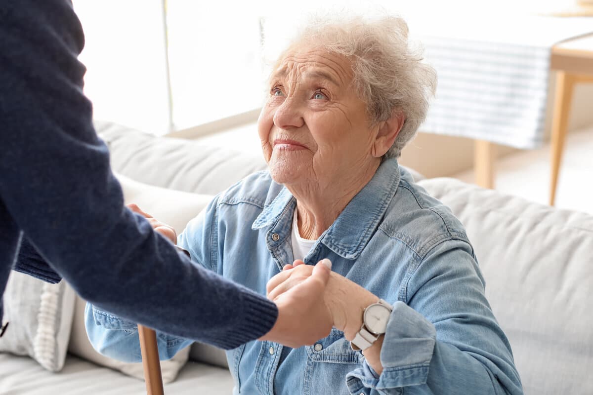 Young caregiver helping senior woman to stand up at home