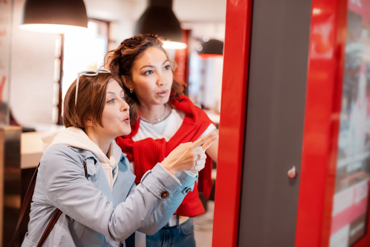 Two girl friends orders food and lunch at a fast food restaurant using a self-service kiosk or a terminal with a screen.