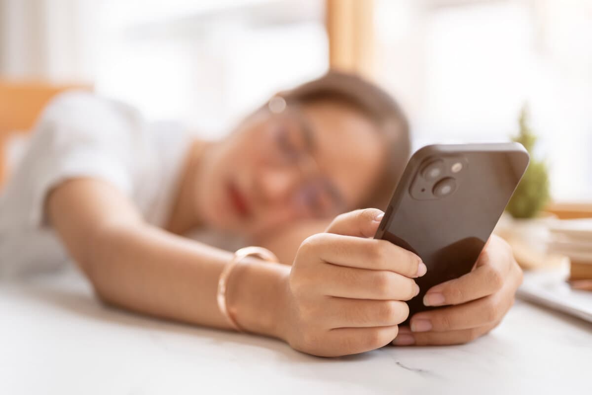 A tired, bored Asian woman leaning on the table and holding her smartphone, waiting for the messages