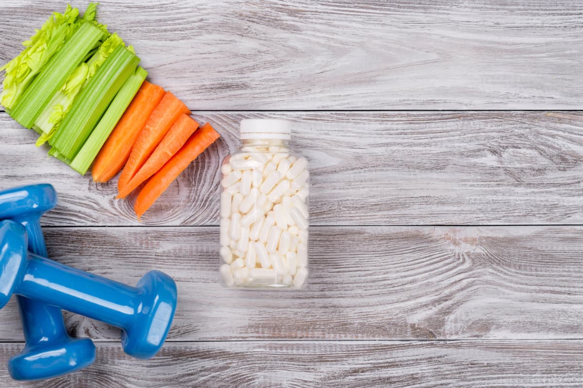 Frühlings-Flatlay-Sportkomposition mit Sellerie, Karotten, blauen Hanteln und L-Carnitin-Vitaminen in Kapseln auf grauem Holzhintergrund. Konzept gesunder Lebensstil, Sport und Ernährung im Frühling. Ansicht von oben.