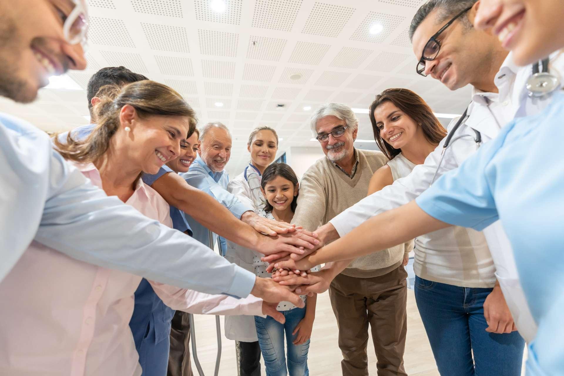 Gruppe von Gesundheitspersonal und Patienten unterschiedlichen Alters in einer Gruppe, alle mit Händen beim Lächeln im Krankenhaus