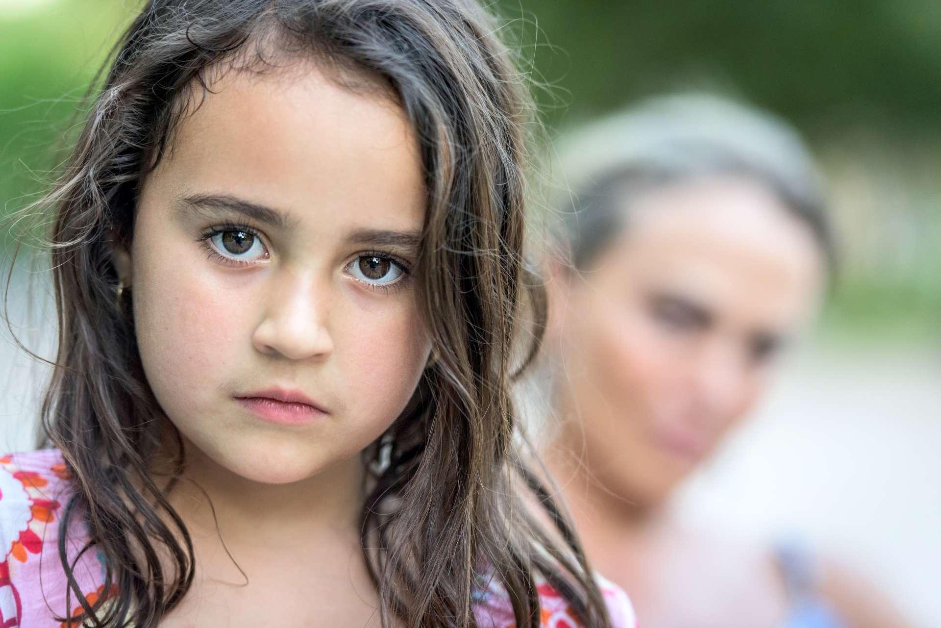 Outdoor-Bild eines ernsten latino sechs Jahre alten kleinen Mädchens posiert mit ihrer Mutter im Hintergrund