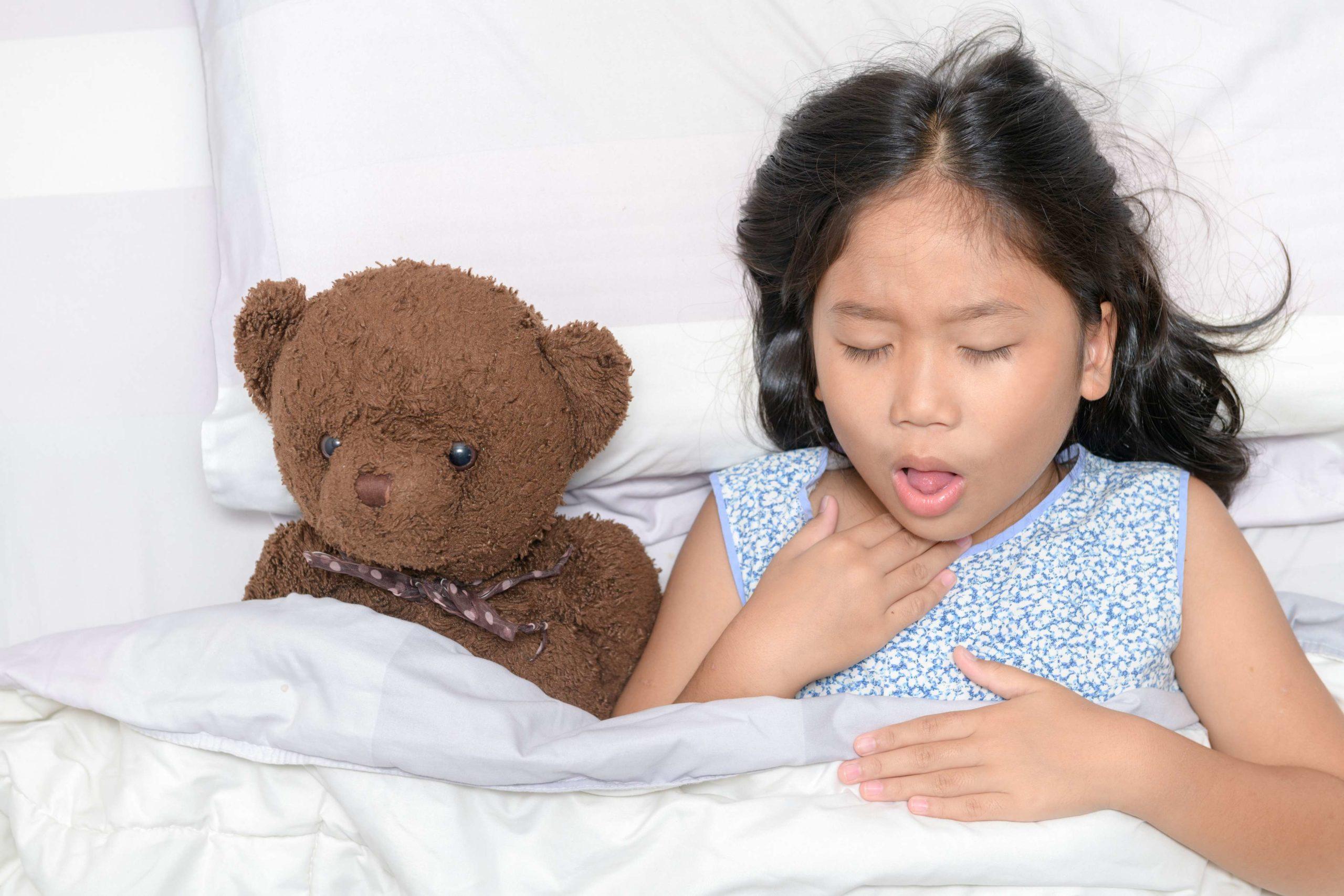 Kleines Mädchen hustet und Halsschmerzen, die auf Bett mit Spielzeugbär liegen, Gesundheitskonzept