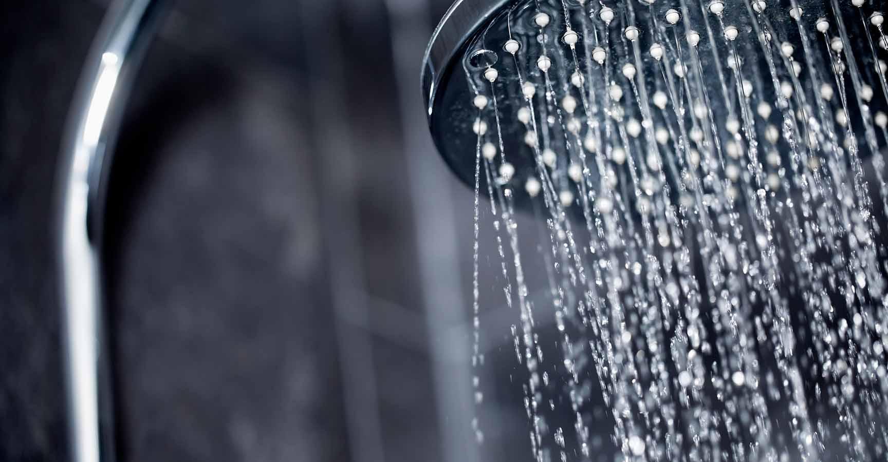 Close up view of large shower head sprinkling water with black tiles in the background.
