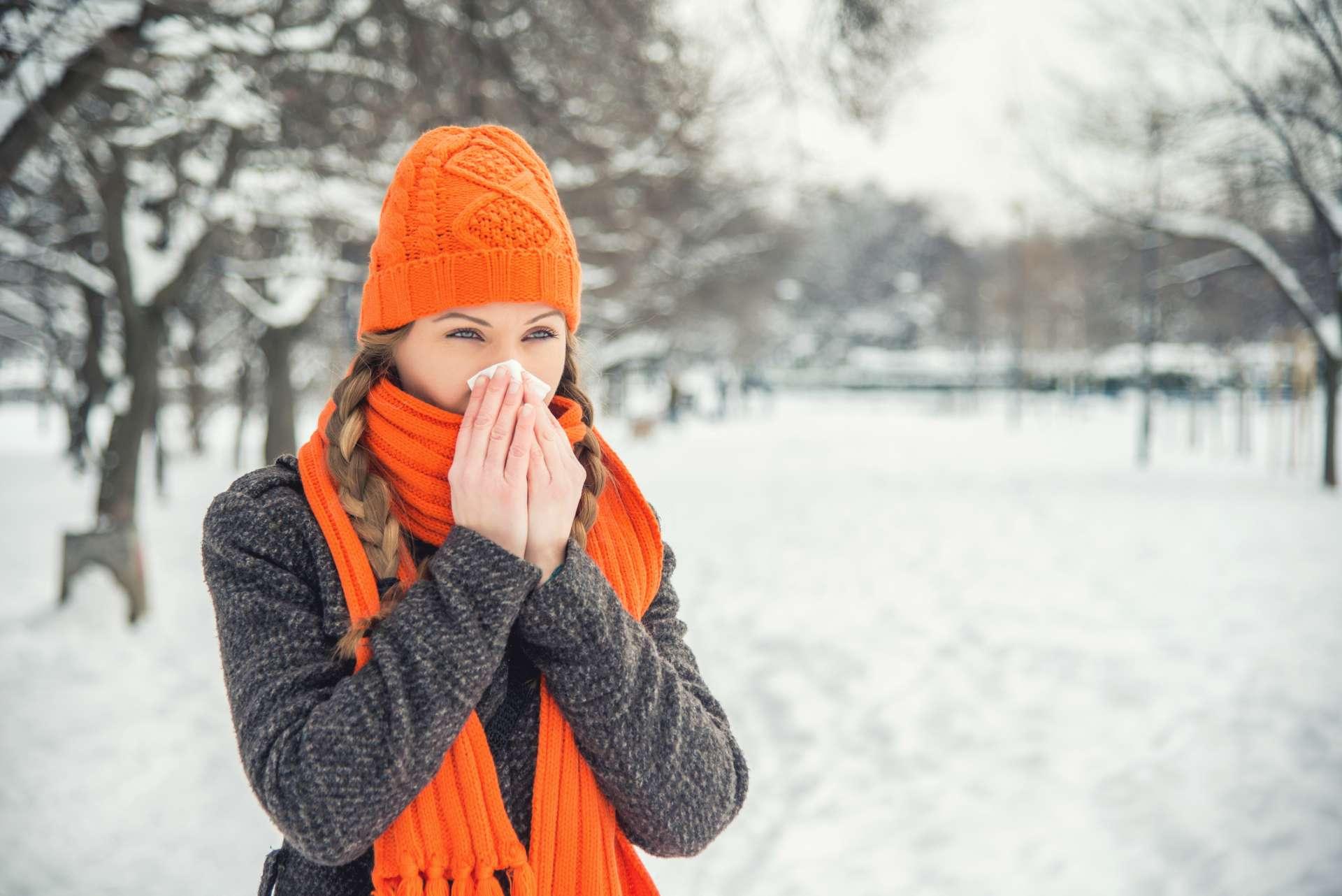 A young woman with braided long blond hair is outdoors in a park during the winter. There is snow and trees in the background. She is wearing winter clothes, an orange hat and scarf, and is blowing her nose with a handkerchief / hanky / tissue. She is looking away from the camera. With copy space.