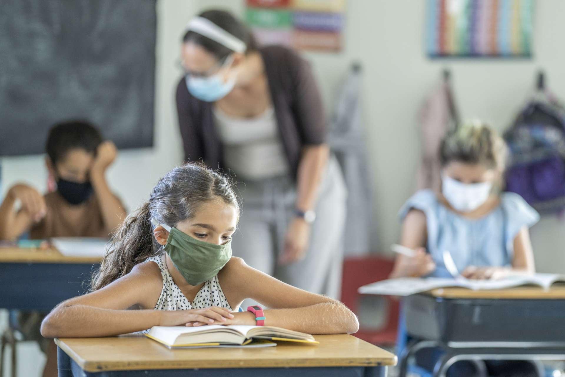 12 Jahre altes Mädchen, das eine wiederverwendbare, schützende Gesichtsmaske im Klassenzimmer trägt, während an der Schularbeit an ihrem Schreibtisch gearbeitet wird.