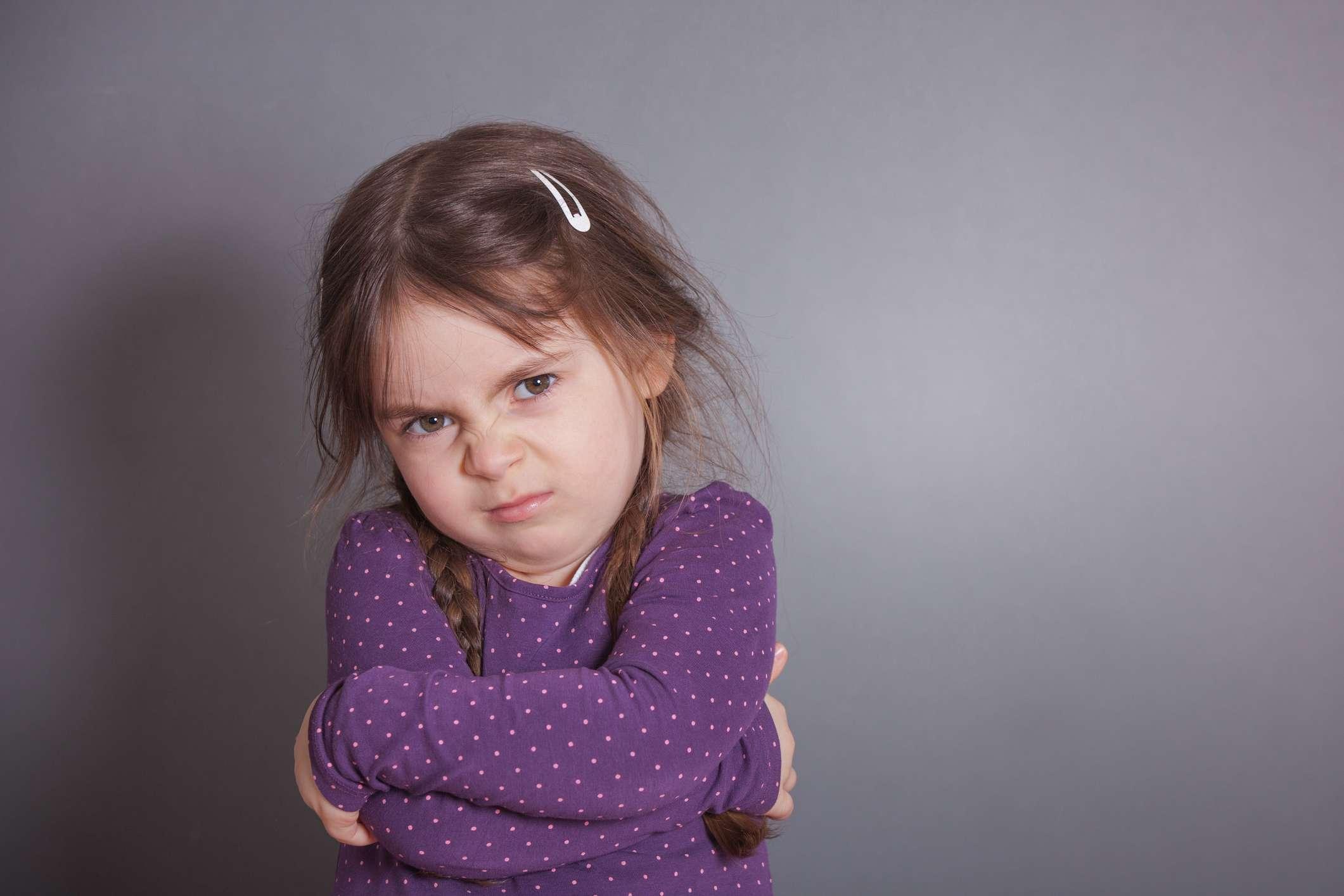 Ein kleines süßes Mädchen schmollt. Sie trägt ein lila Hemd vor einem grauen Hintergrund.