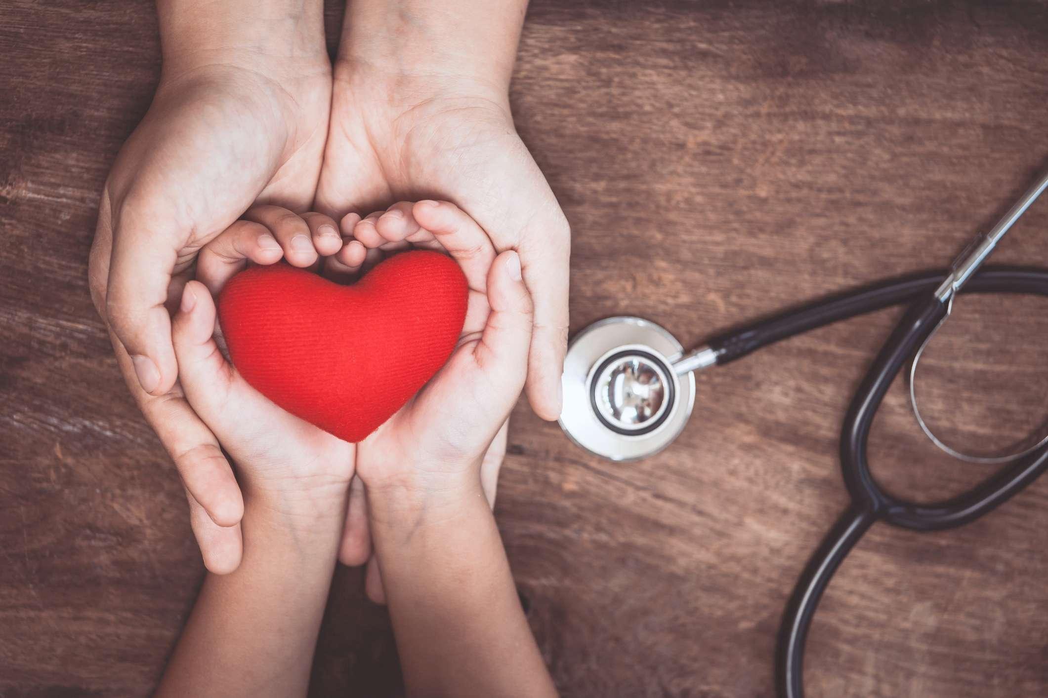 Rotes Herz auf Frauen- und Kinderhänden und mit Stethoskop des Doktors auf hölzernem Hintergrund. Medizinisches Konzept des Gesundheitswesens