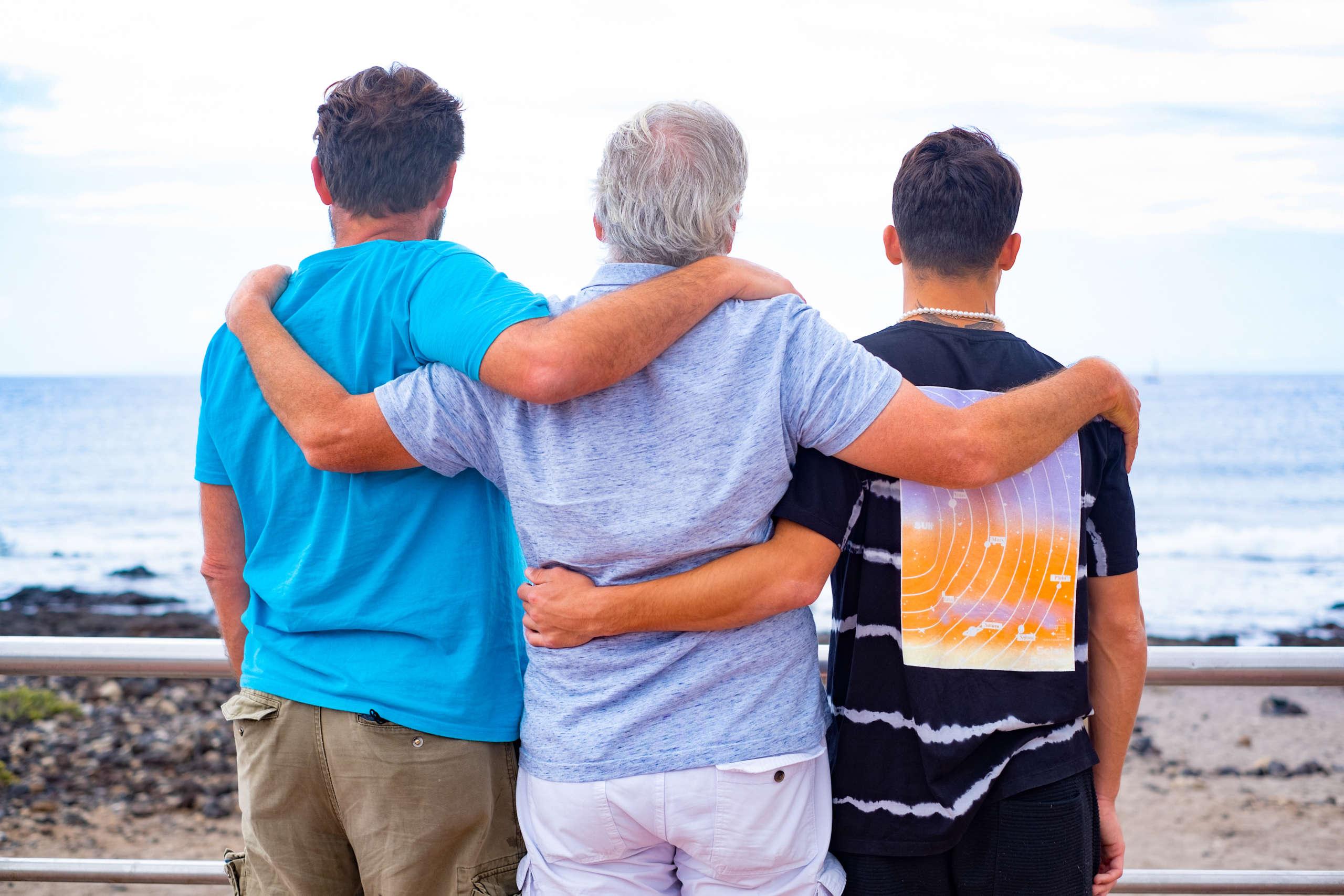 Familiengruppe, drei Männer, Vater, erwachsener Sohn und Teenager-Enkel, die sich umarmen und den Horizont über Wasser betrachten