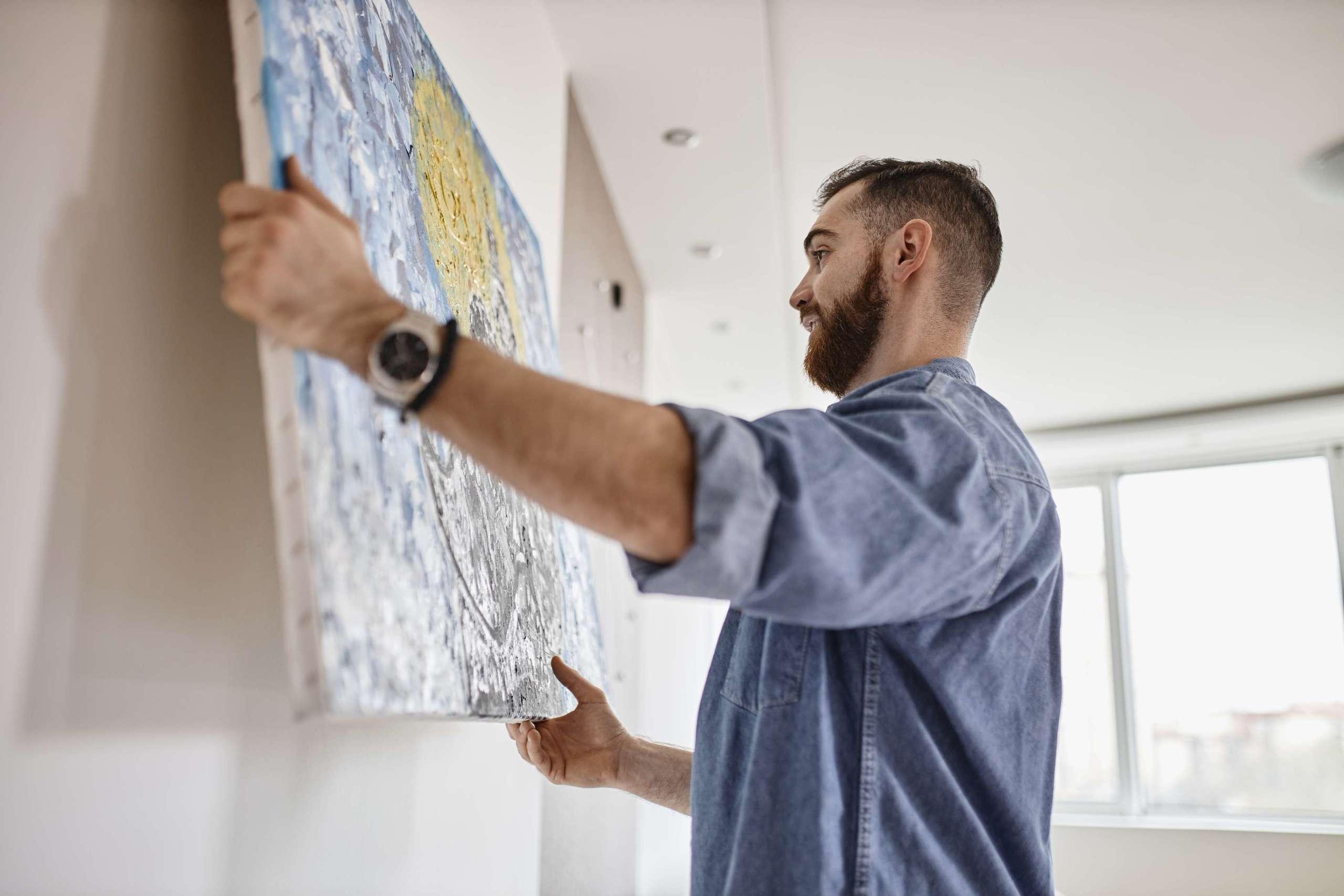 Mann mit Bart, der in seinem Wohnzimmer ein Gemälde an die Wand hängt