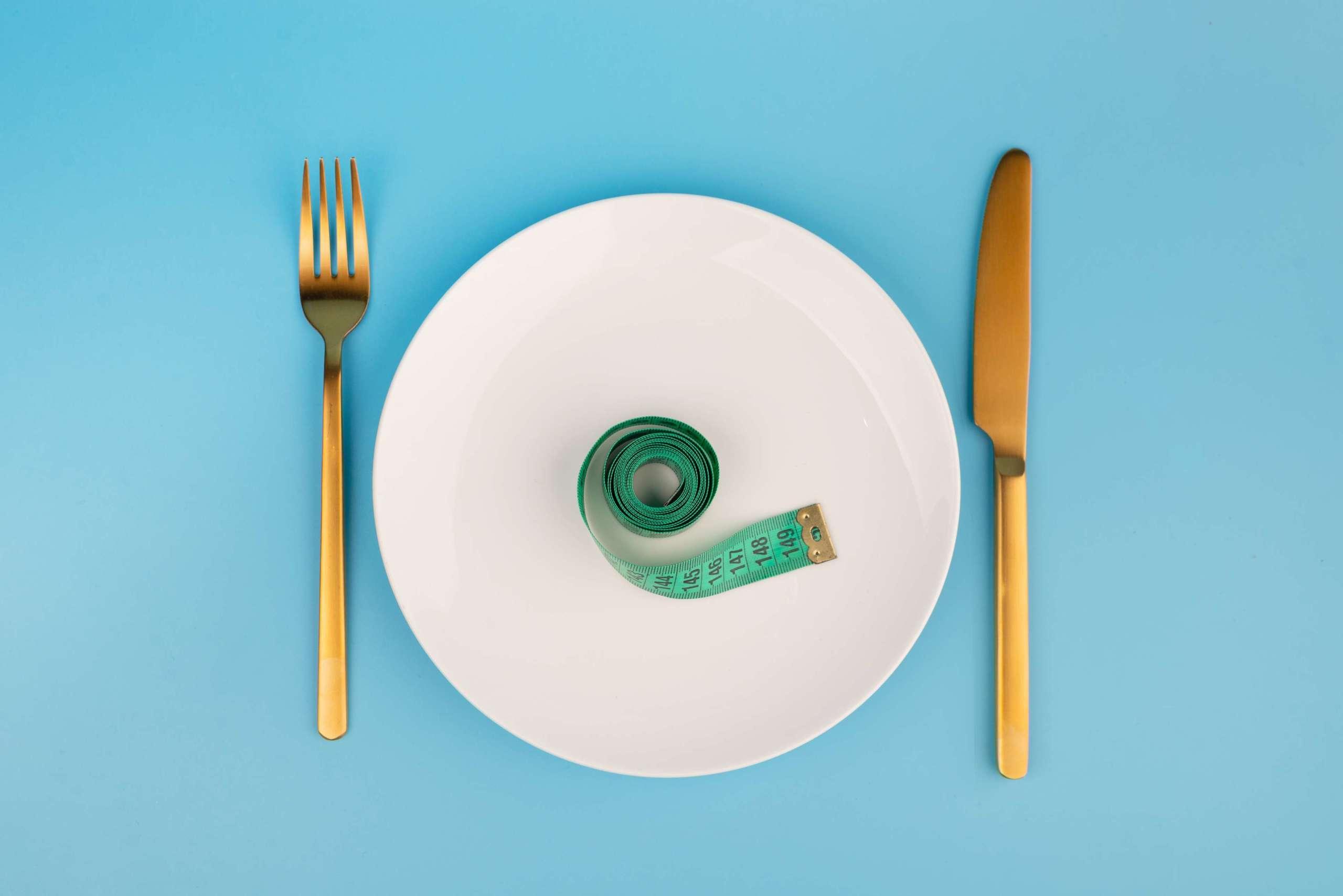 weiße runde Platte und goldene Gabel und Messer mit grünem Maßband auf blauem Hintergrund, Gewichtsverlustkonzept, flache Lage.