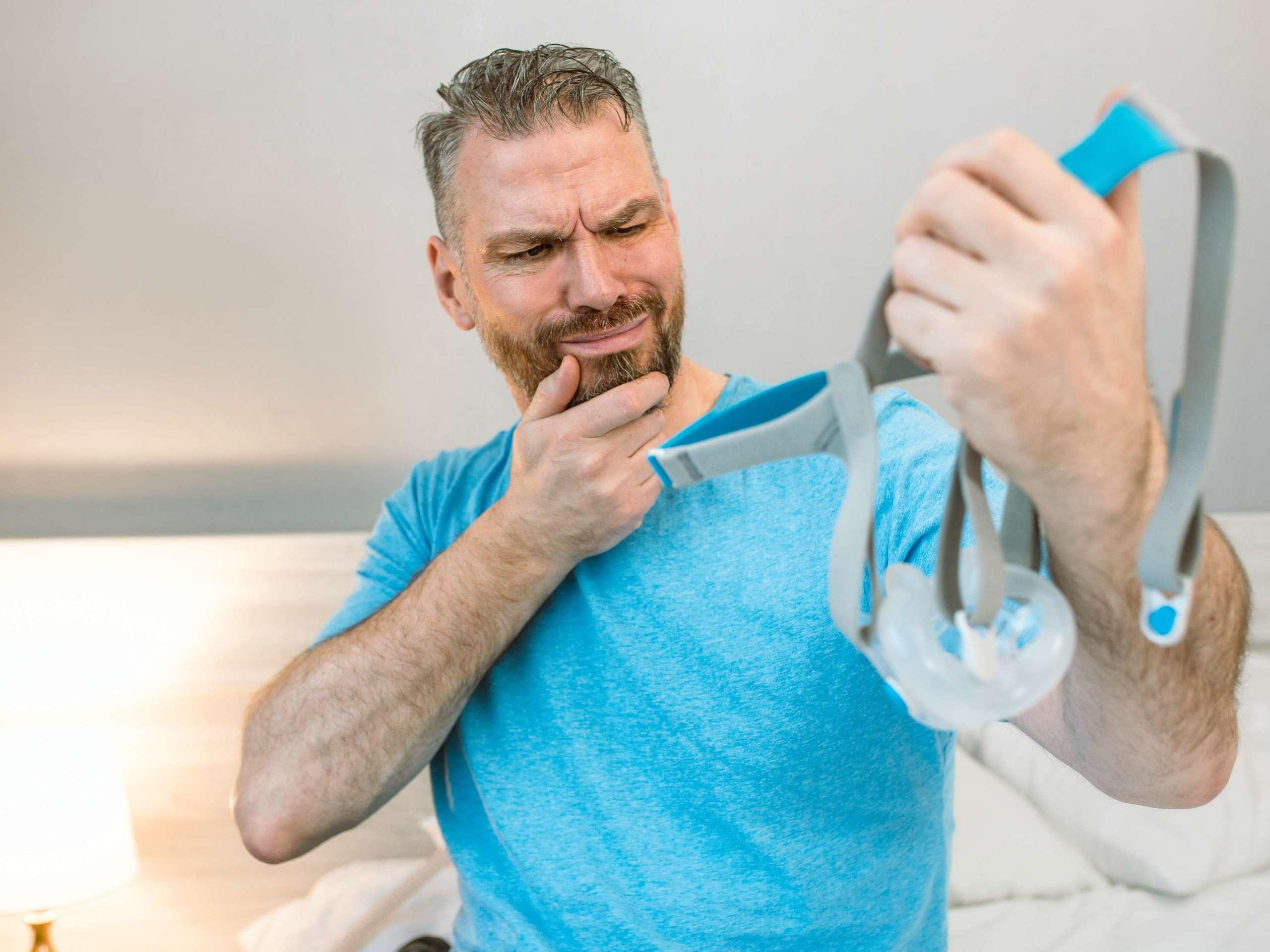 Unglücklicher schockierter Mann mit chronischen Atemproblemen überrascht, indem er ein CPAP-Gerät verwendet, das auf dem Bett im Schlafzimmer sitzt.