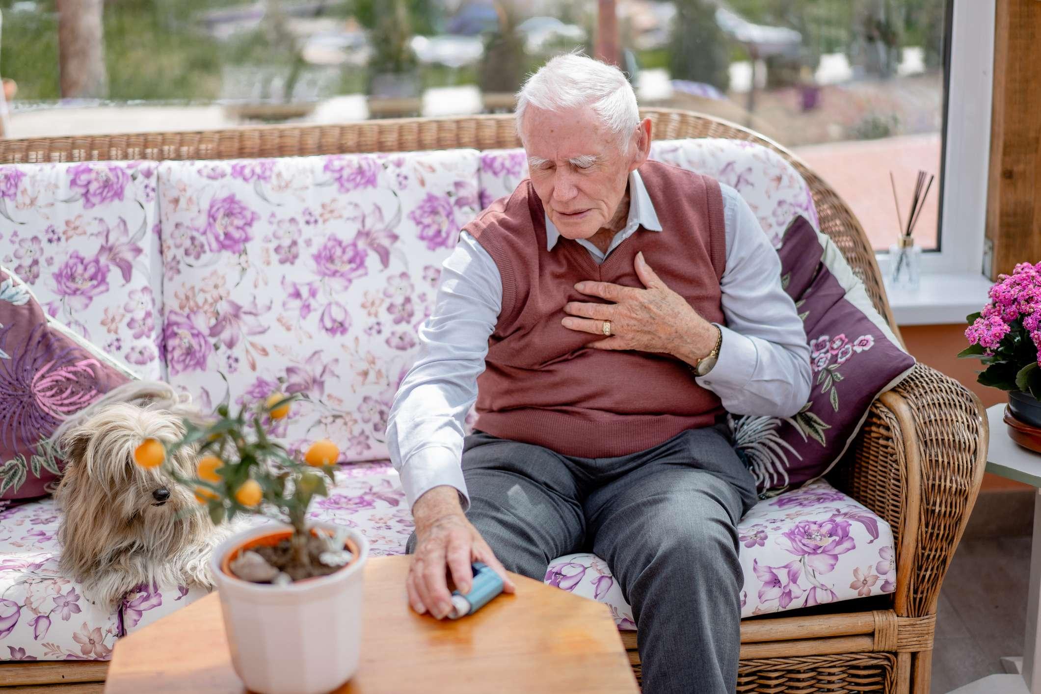 Ein älterer Mann, der zu Hause einen Asthmaanfall hat, während er auf seinem Sofa sitzt. Er bereitet sich auf die Inhalation vor
