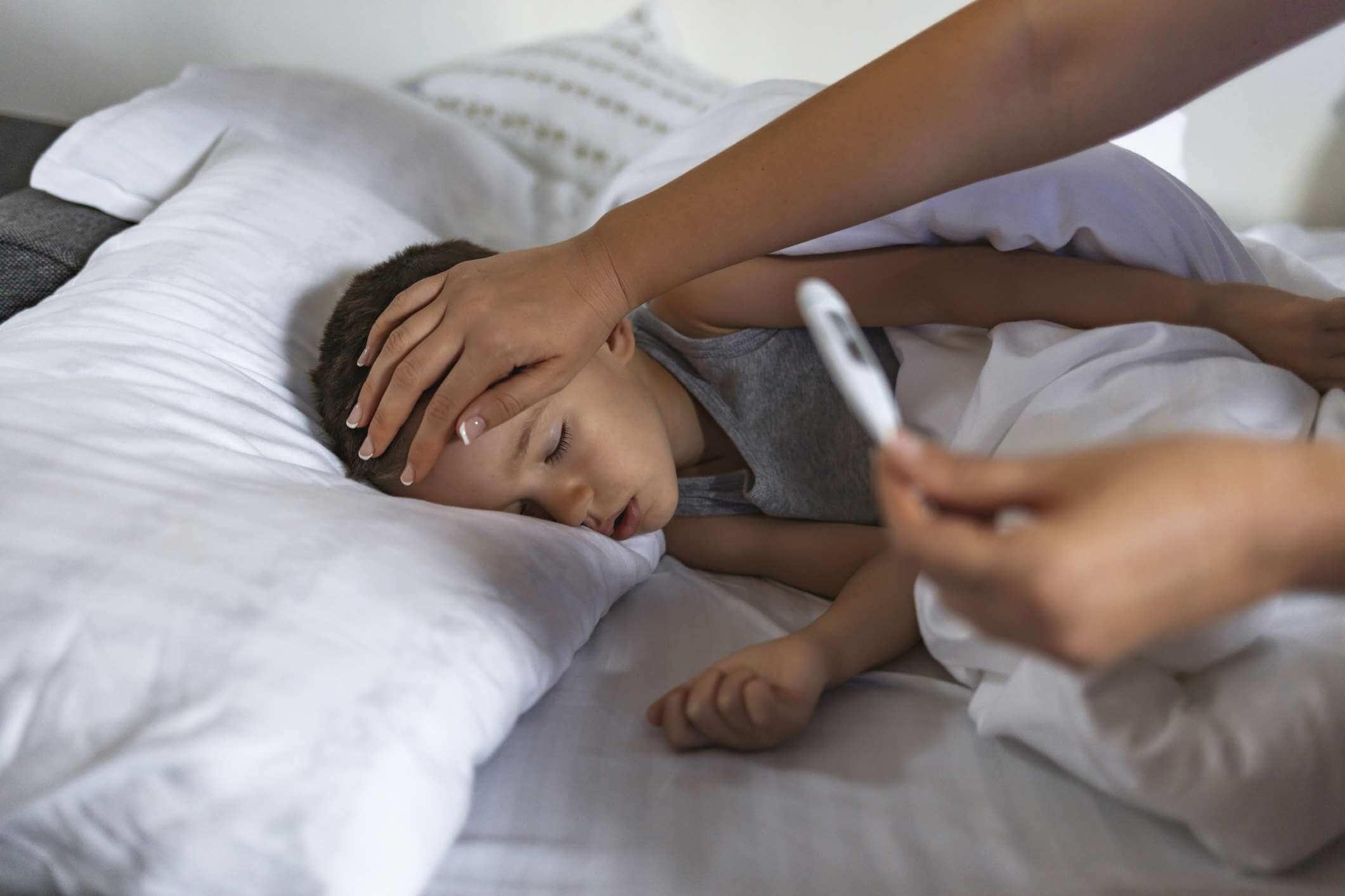 Foto eines kranken männlichen Kindes mit hohem Fieber, das im Bett liegt, während die Mutter die Temperatur misst.