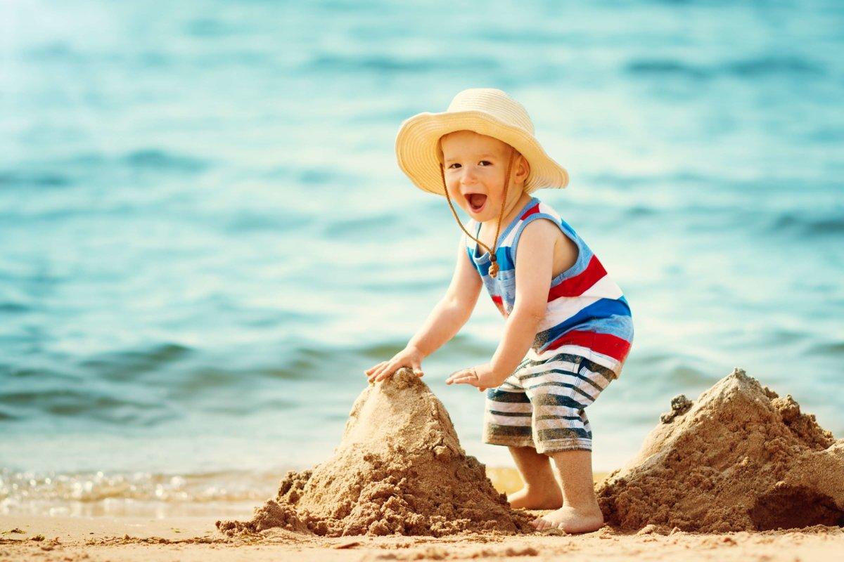 Einjähriger Junge, der mit Strohhut am Strand spaziert. Kind im Familienurlaub am Meer beim Bau einer Burg