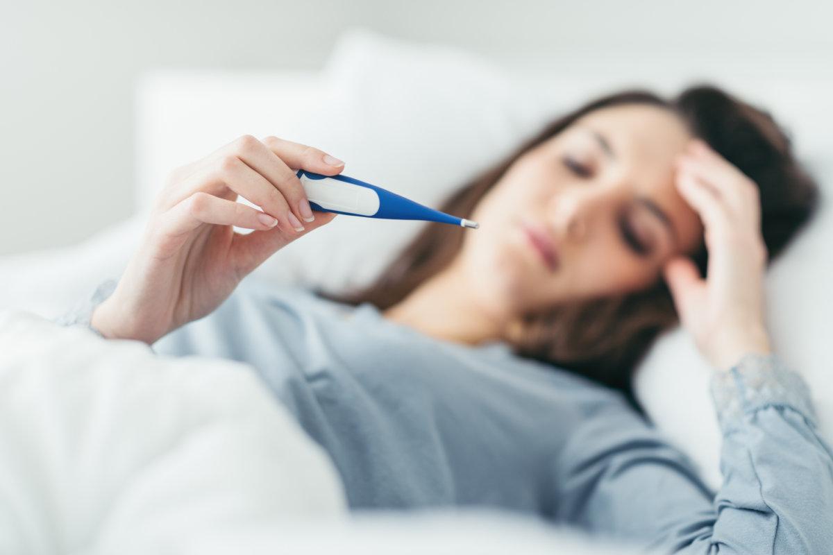 Frau mit Grippevirus liegt im Bett, sie misst ihre Temperatur mit einem Thermometer und berührt ihre Stirn