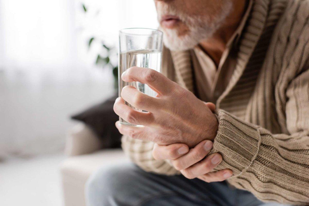cropped view of aged man with parkinsonism holding glass of water in trembling hands while sitting at home,stock image